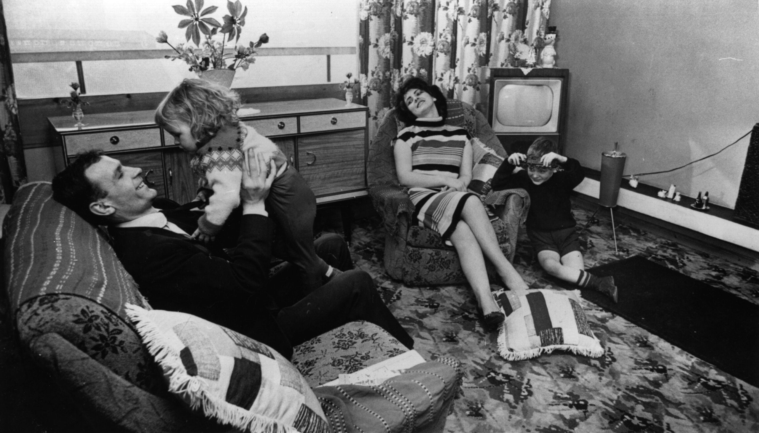 A man plays with a small child on a sofa while a woman sits in an armchair and another child plays on the floor in a living room with retro decor and a television. Family life in the 1960s and 70s.