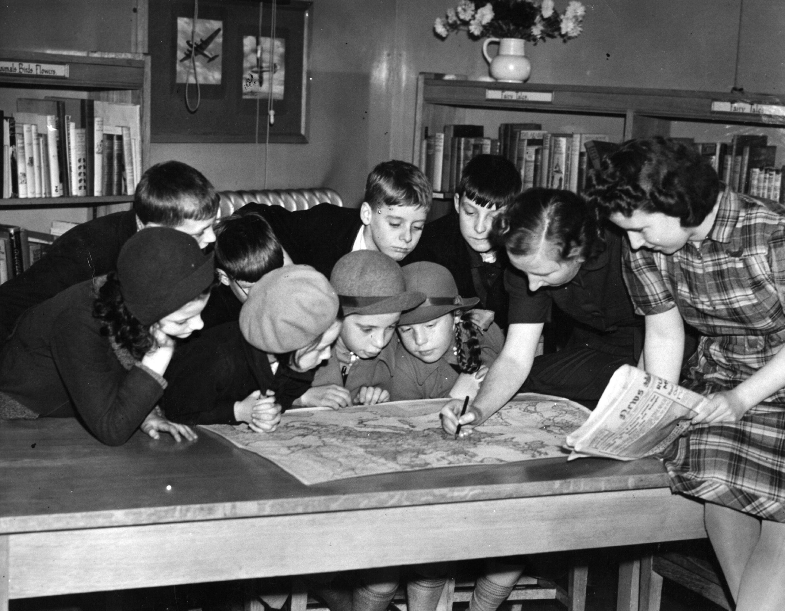 A group of children gather around a table, looking at a map while a woman points at it. Bookshelves and a vase of flowers are in the background. One child holds a newspaper.
