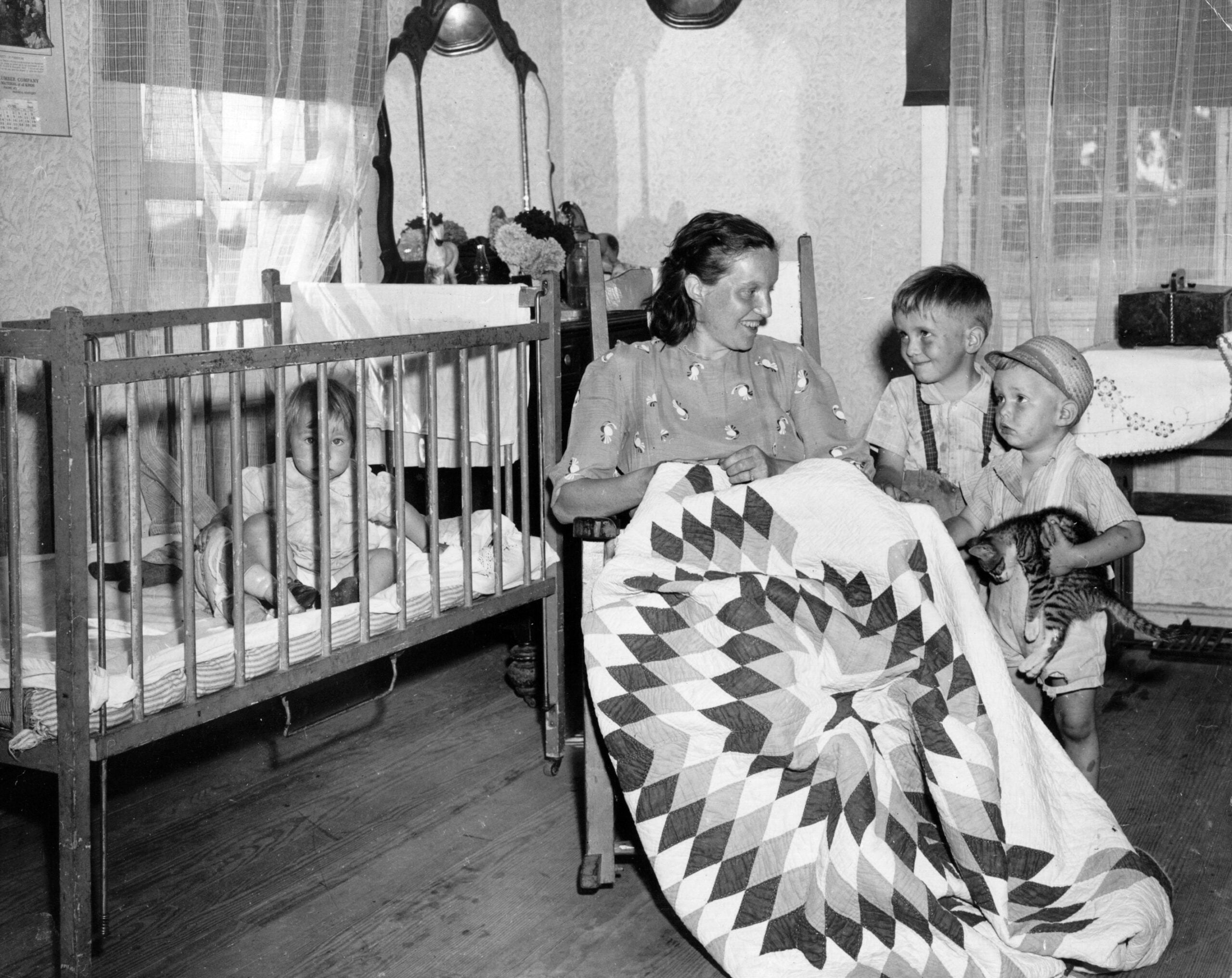 A woman sits with two young boys and a cat beside a crib with a toddler inside. The woman holds a patterned quilt. The room has wooden floors, lace curtains, and furniture in the background.