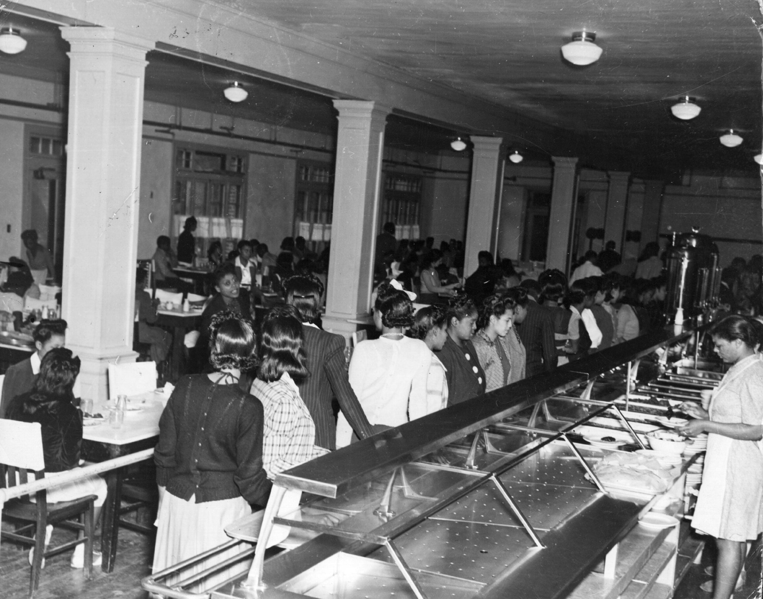Black and white photo of people standing in line at a cafeteria counter, with others seated at tables in the background. The room has columns and ceiling lights. A woman serves food behind the counter.
