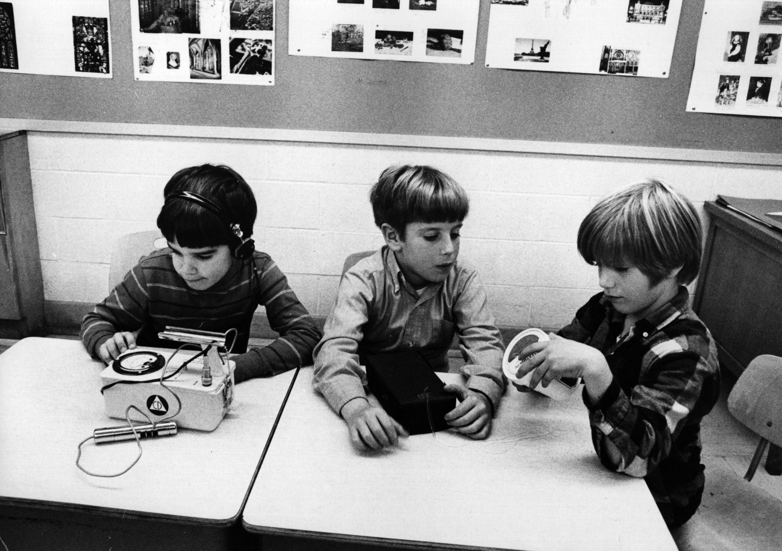 Three young boys sit at desks in a classroom, each using different audio equipment. One wears headphones, another examines a device, and the third holds a reel of tape. Classroom bulletin boards are visible in the background.