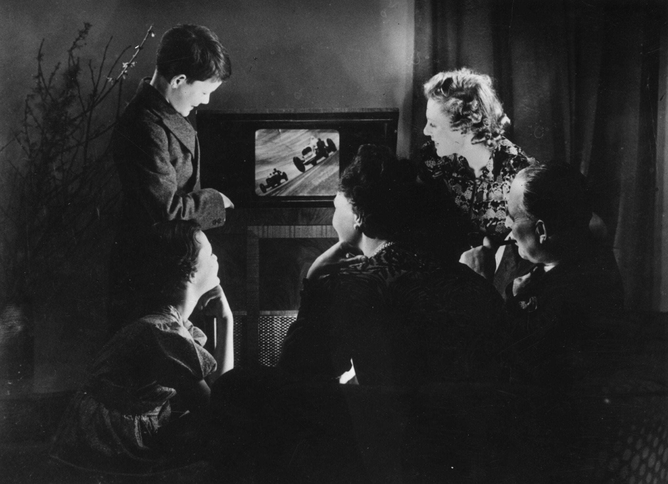 Five people sit and stand around a vintage television, watching a black-and-white program. The scene is indoors at night, with dim lighting and curtains in the background.