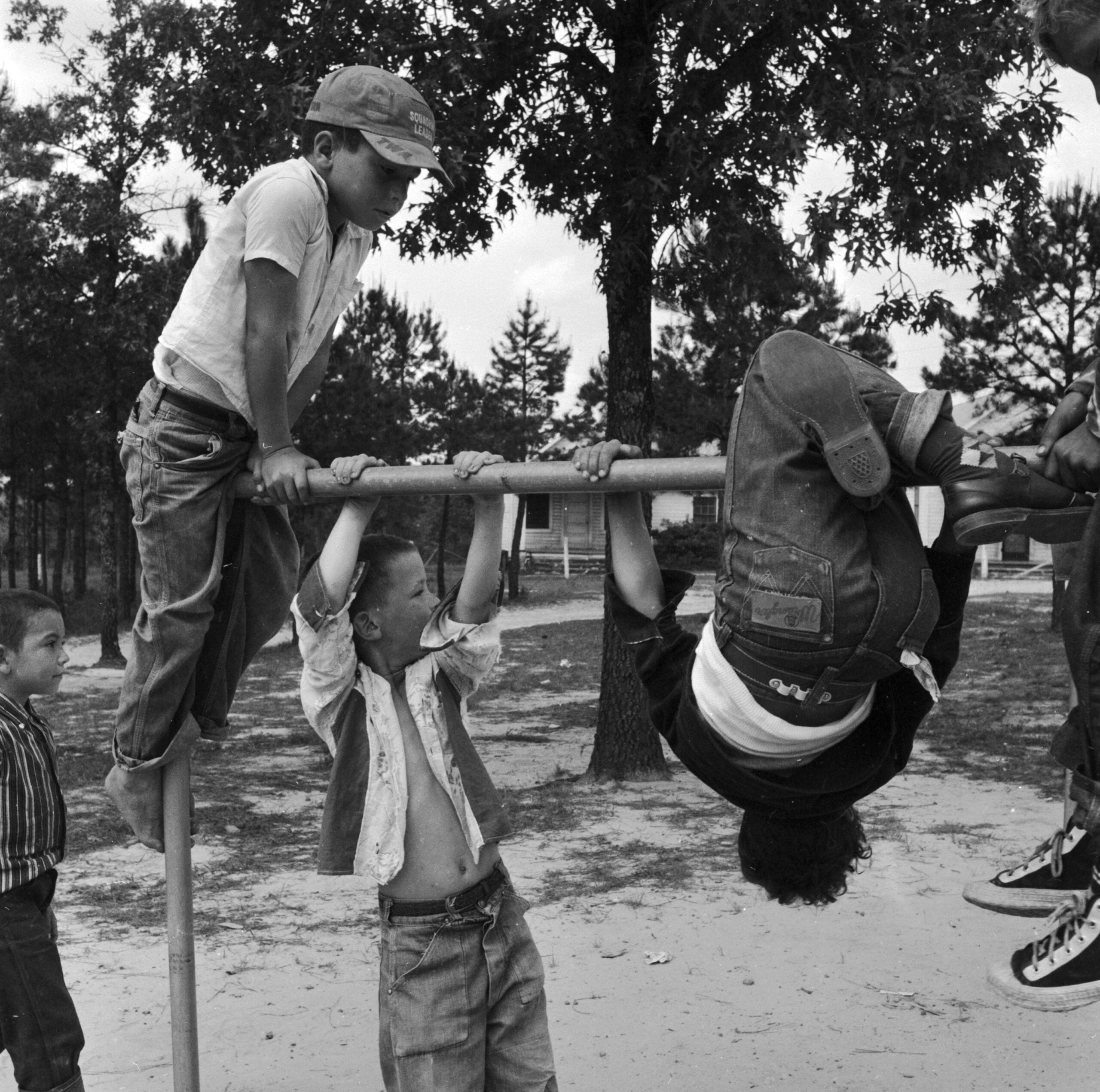 Four children play outside on a horizontal bar; one hangs upside down while two others hold onto the bar. Trees and a small building are visible in the background.