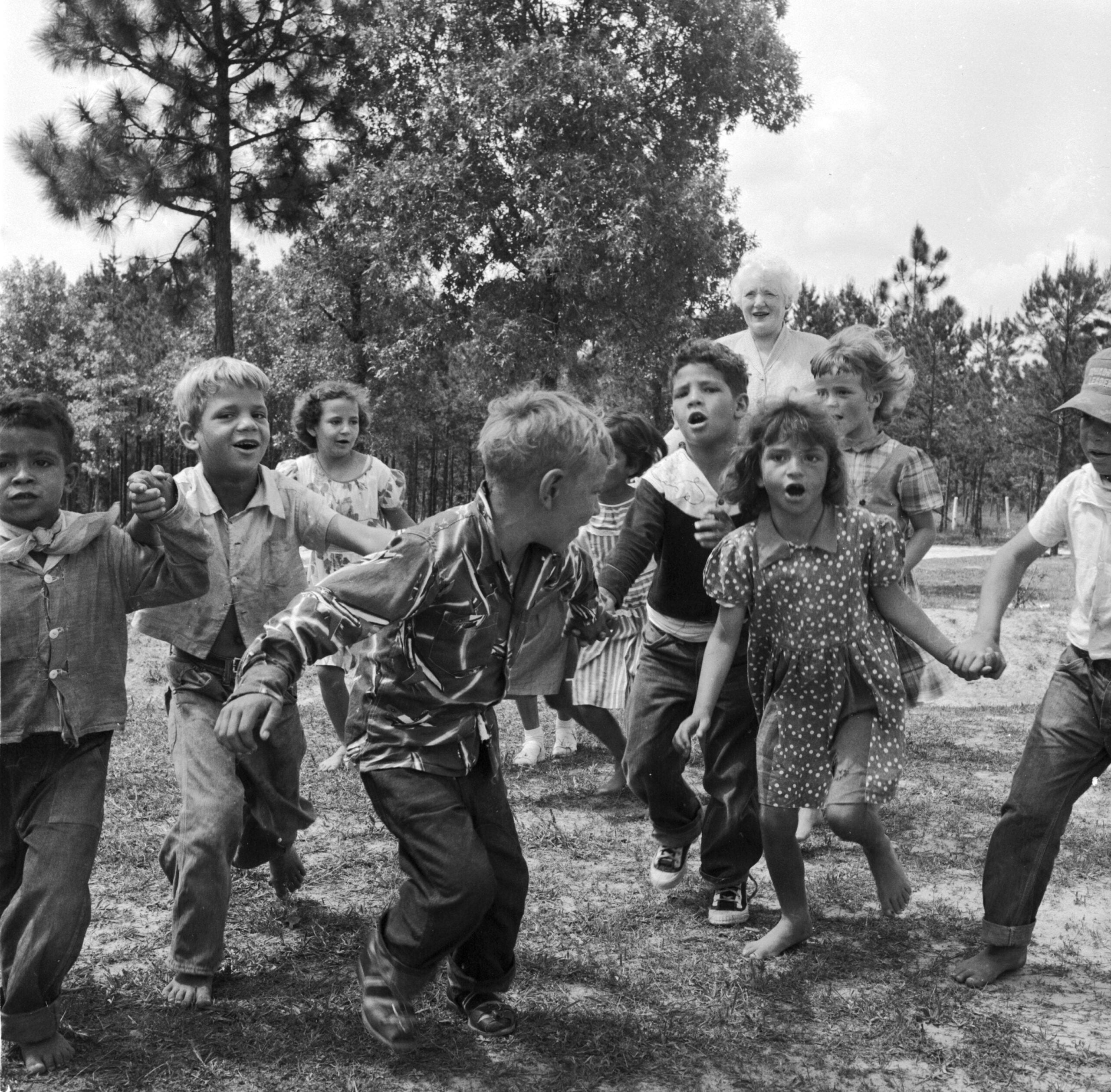 A group of children holding hands and playing outside on a grassy field, surrounded by trees. Some children appear to be mid-jump, and everyone looks happy and energetic.