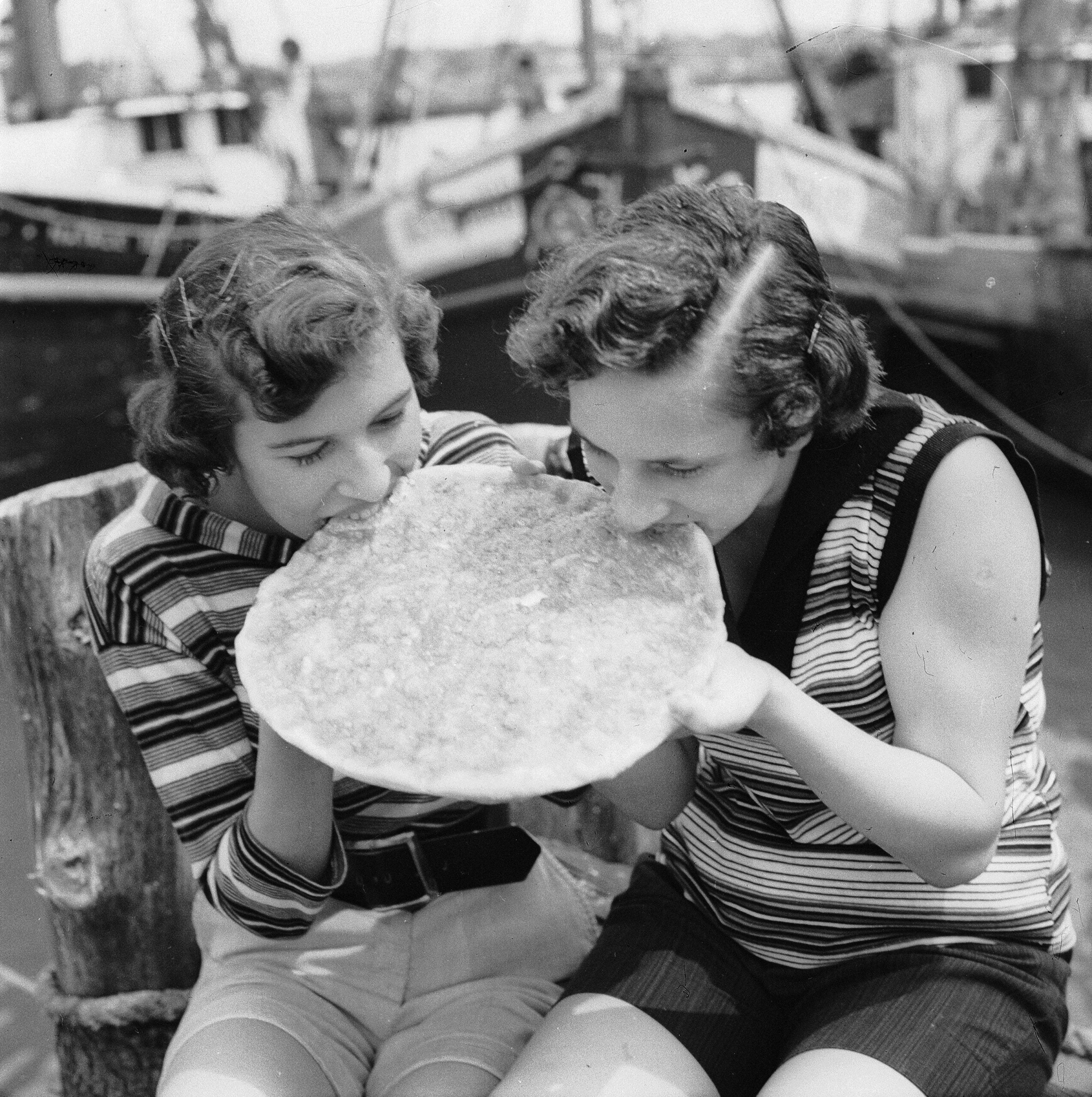 Two women in striped tops sit side by side outdoors, both biting into a large, flat piece of food, possibly bread or pastry, near a docked boat in the background.