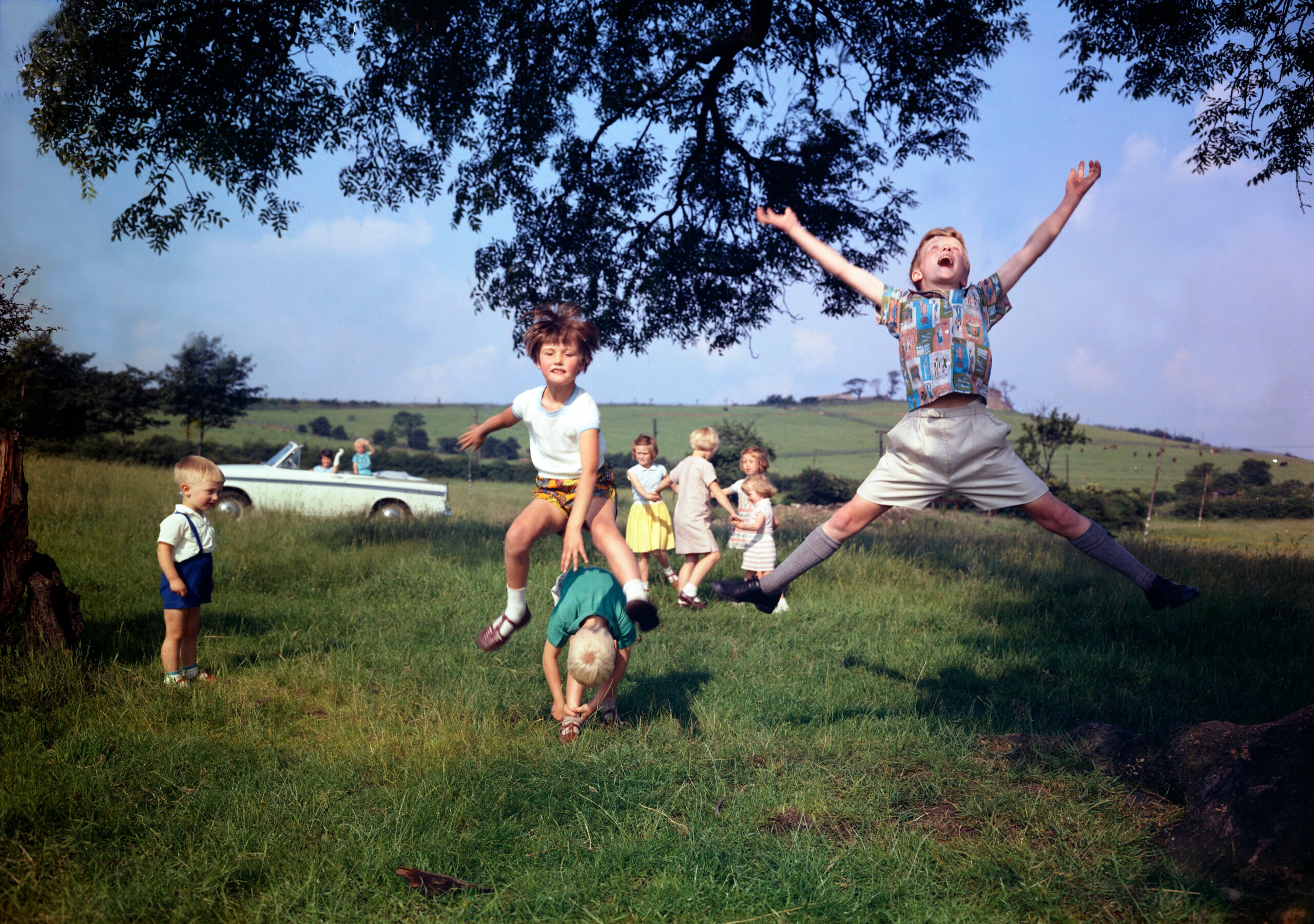Children playing outdoors in a grassy field, some jumping energetically, with a classic white car and rolling hills in the background on a sunny day.
