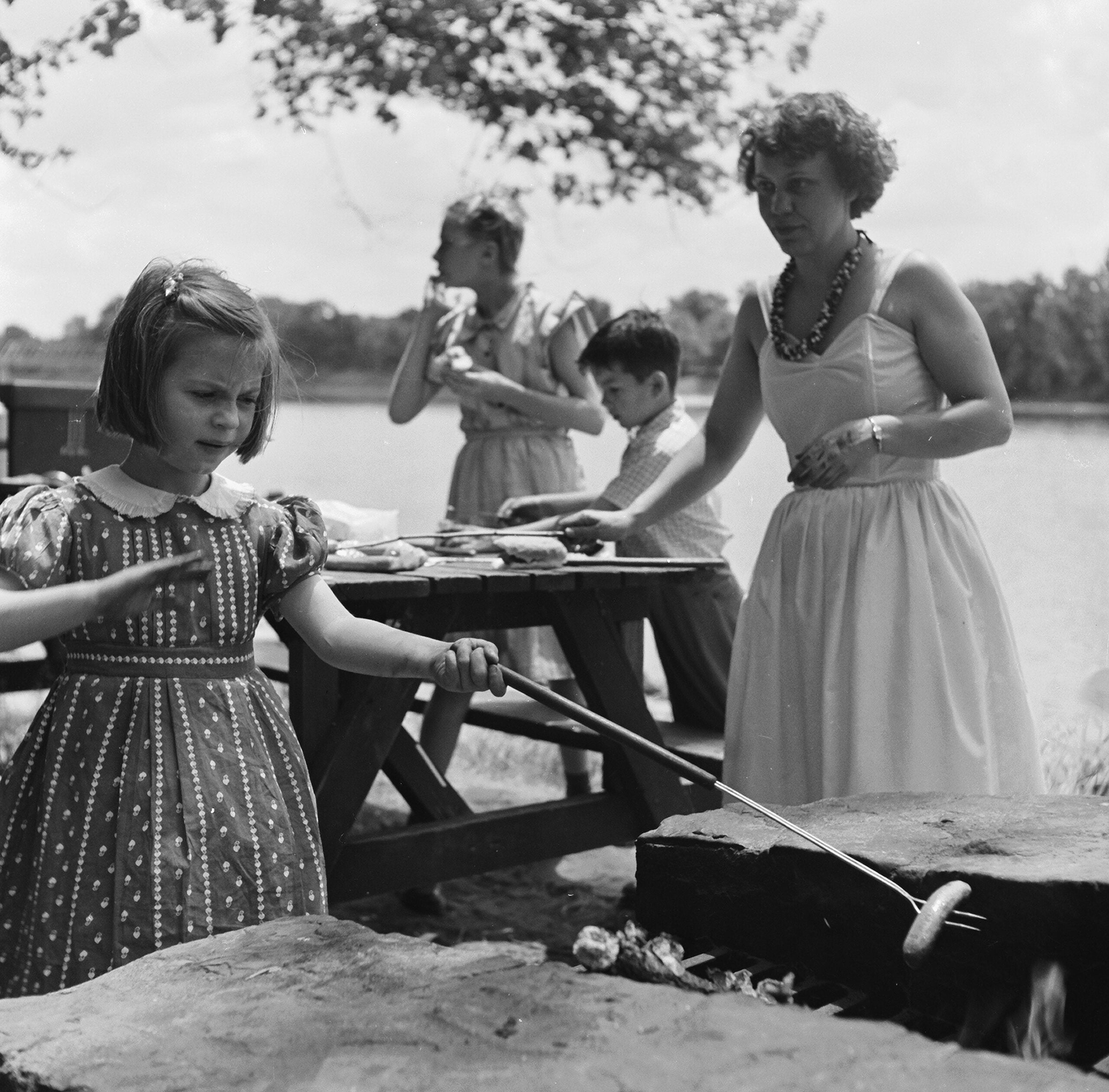 A young girl roasts a hot dog over a fire pit while a woman and two children prepare food at a picnic table near a lake on a sunny day.