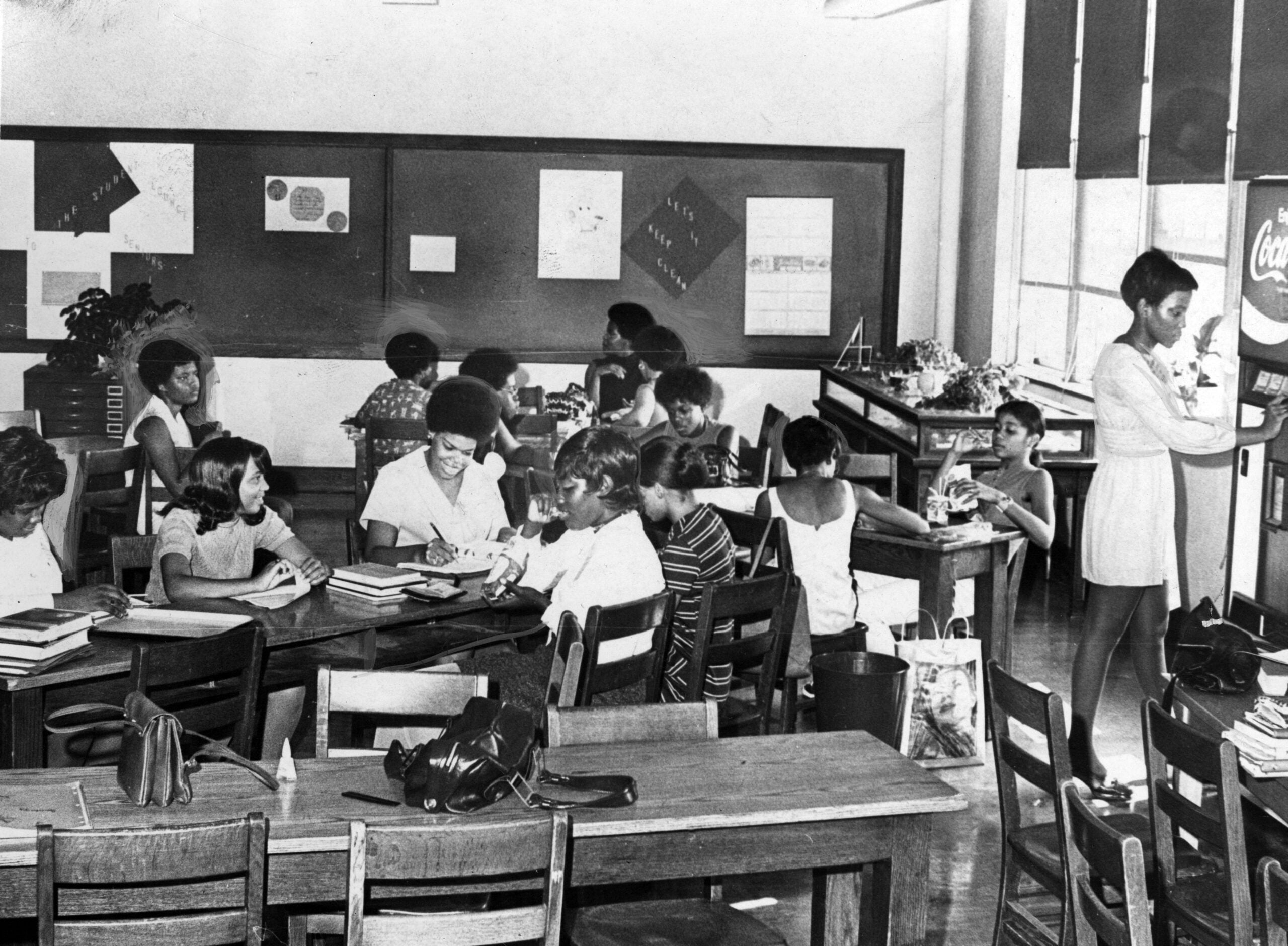 A group of young girls sit at tables in a classroom, reading and writing. One girl stands at a vending machine. The classroom has large windows, bulletin boards, and books on the tables.