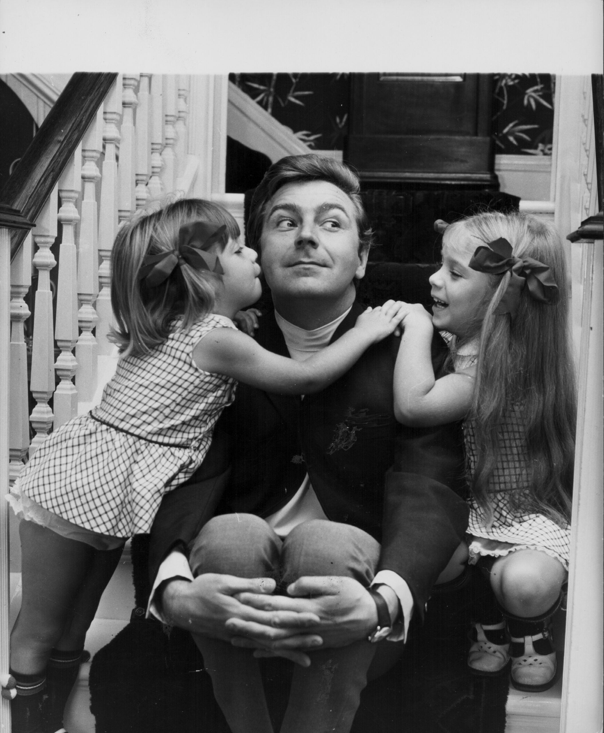 A man sits on stairs, smiling as two young girls with bows in their hair, one on each side, lean in affectionately toward him.