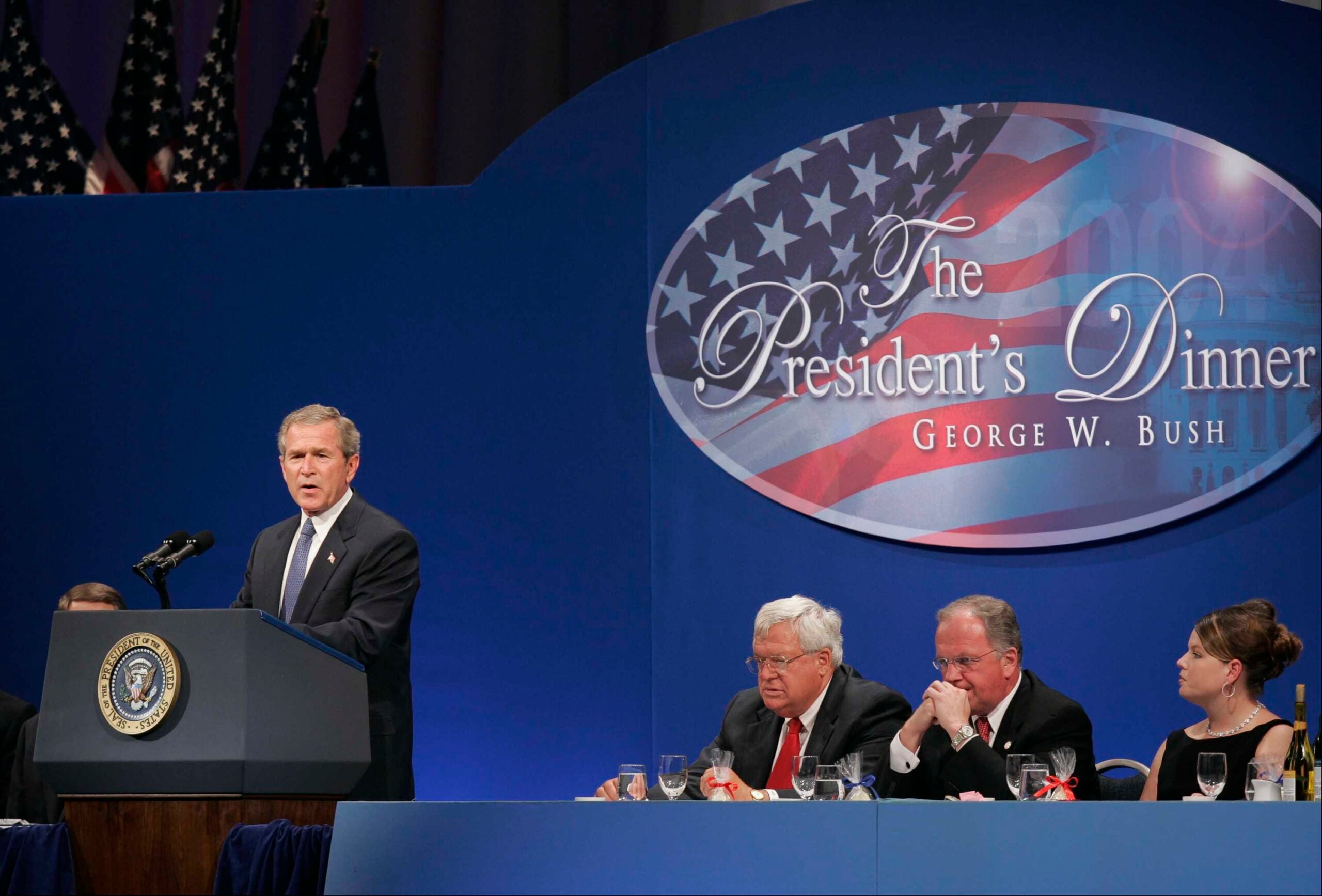 A man stands at a podium with the U.S. presidential seal, addressing a formal dinner. Three men and one woman sit at a table beside him under a sign reading “The President’s Dinner George W. Bush.”