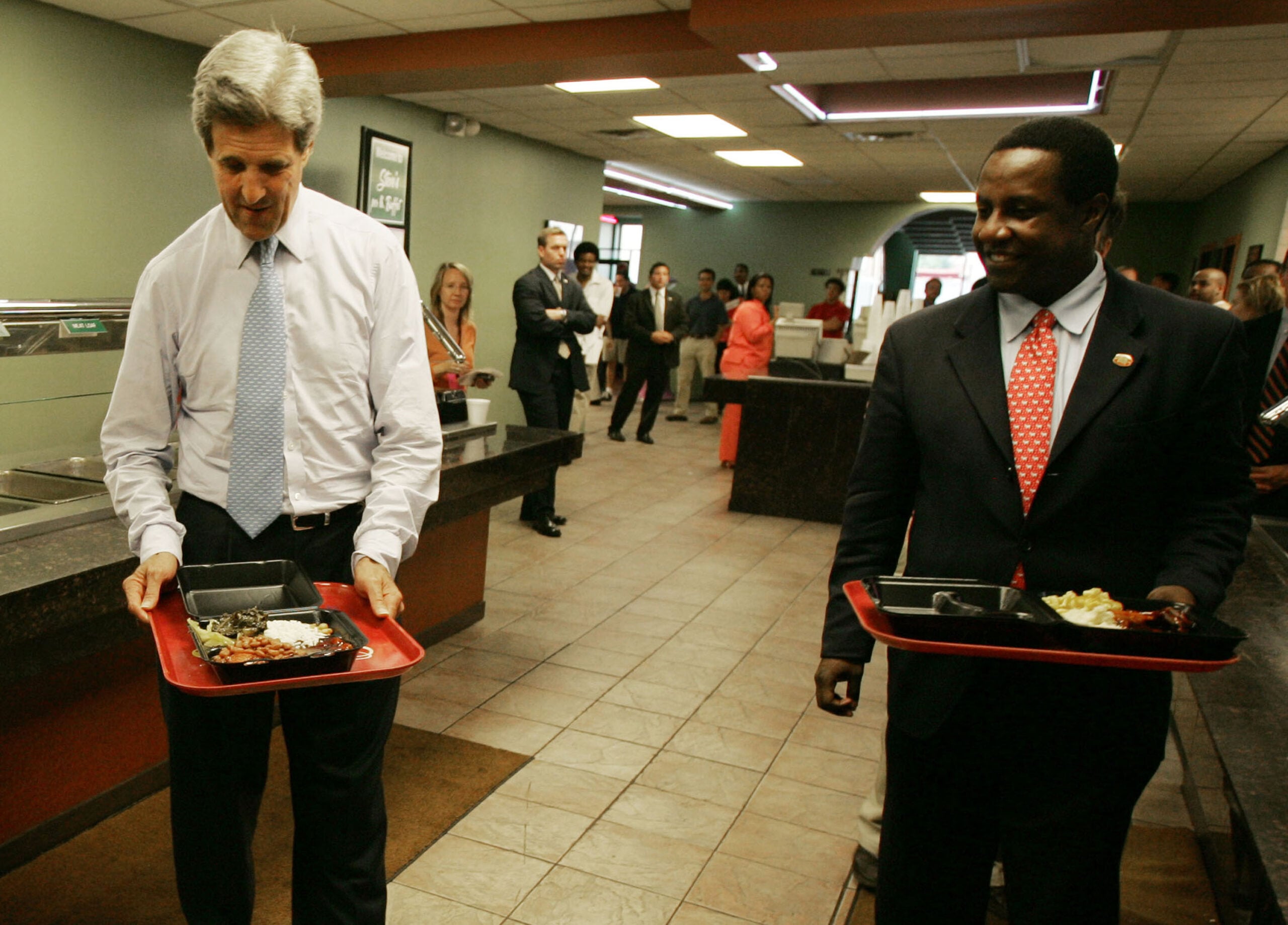 Two men in suits carrying lunch trays walk through a cafeteria line, with other people standing and watching in the background. Both men appear to be engaged in conversation or interaction.