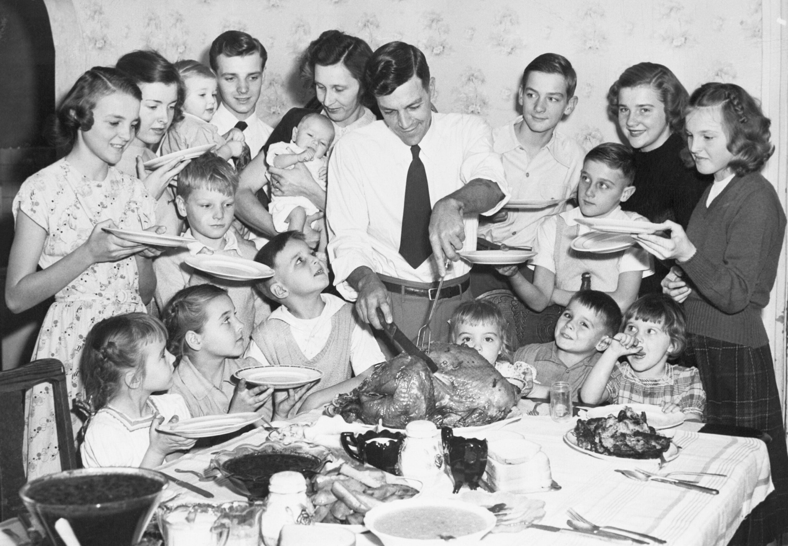 A large family gathers around a dinner table as a man carves a turkey. Children and adults hold plates, eagerly waiting for food. The atmosphere is joyful and festive.