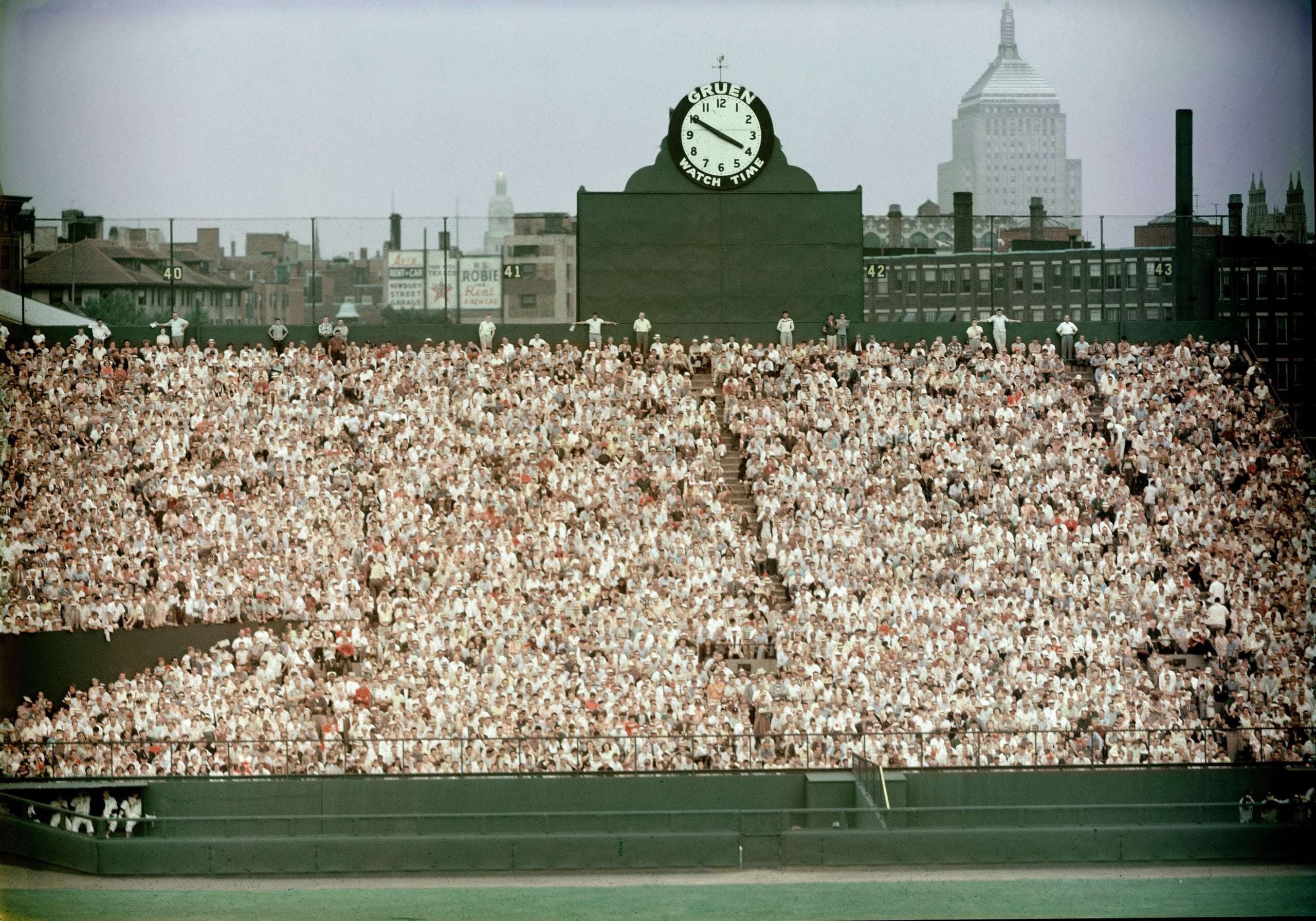 A packed baseball stadium outfield with a large clock above the crowd, city buildings and the Empire State Building visible in the background.