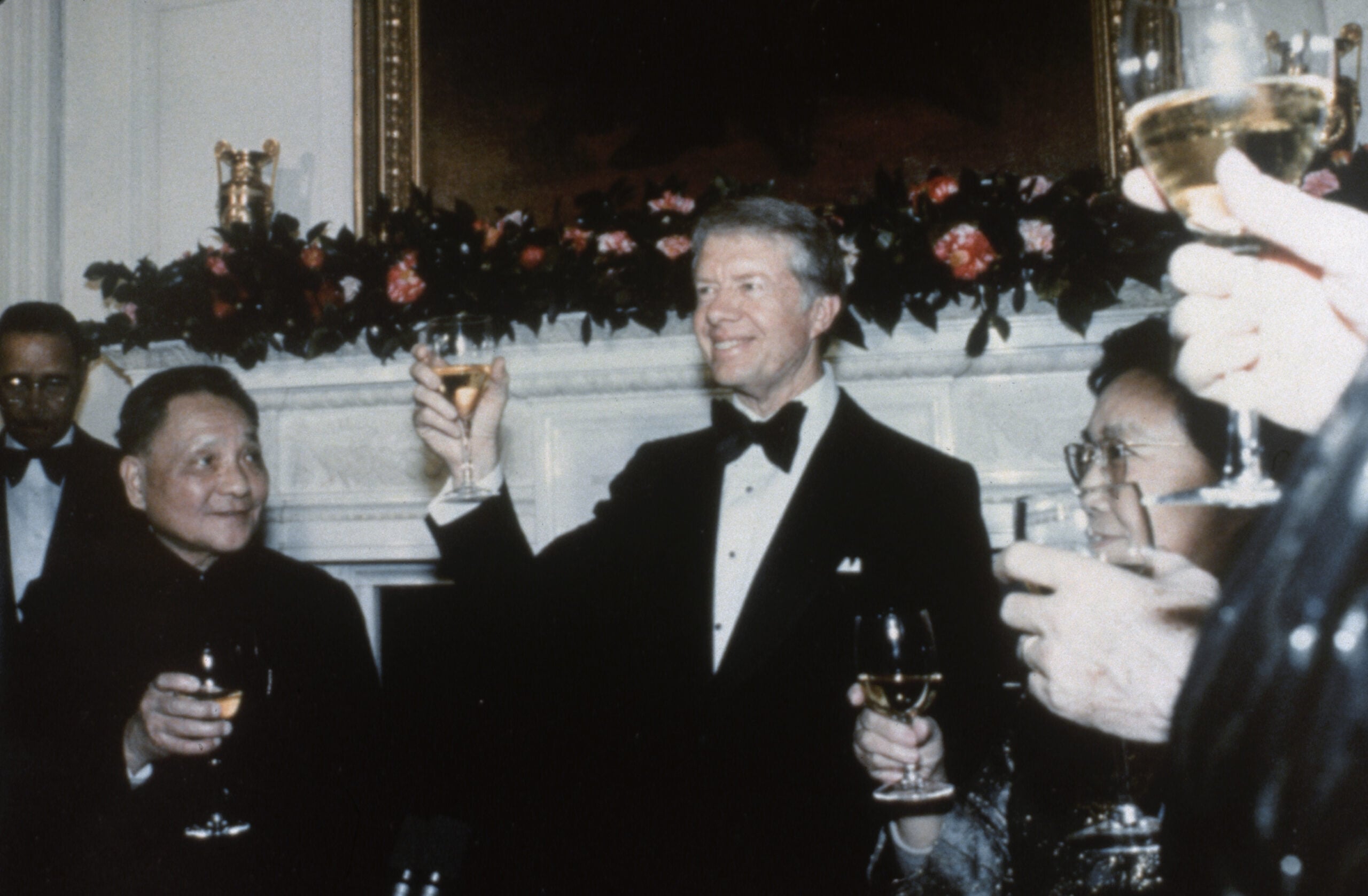 A man in a tuxedo raises a glass in a toast at a formal event, surrounded by others holding drinks, with flowers and a painting in the background.