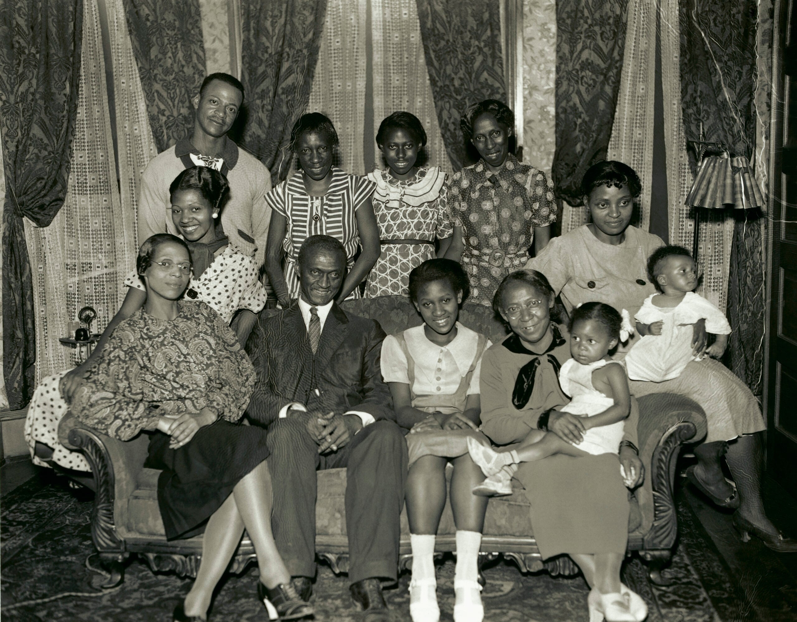 A Black family poses indoors for a formal portrait; three men, seven women, and two young children are seated and standing in front of curtained windows, all dressed in 1930s clothing, looking at the camera.