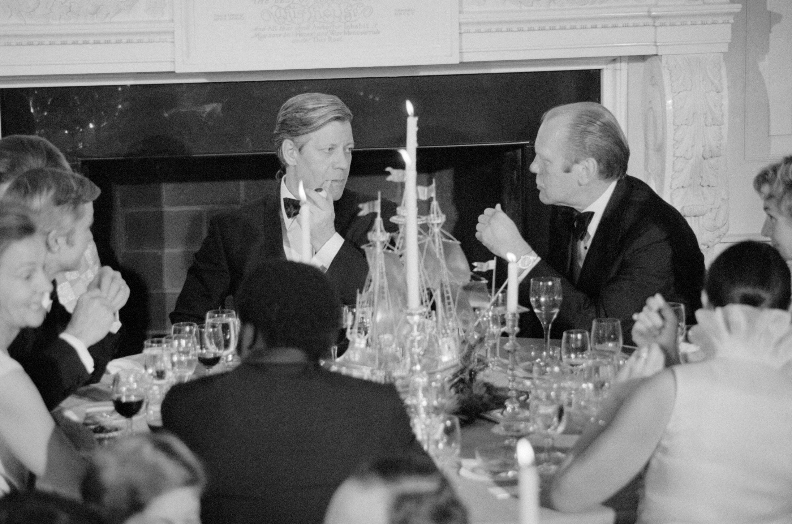 A formal dinner scene with men in tuxedos and women in elegant attire seated around a candlelit table, engaged in conversation. Ornate decorations and glassware are visible on the table.