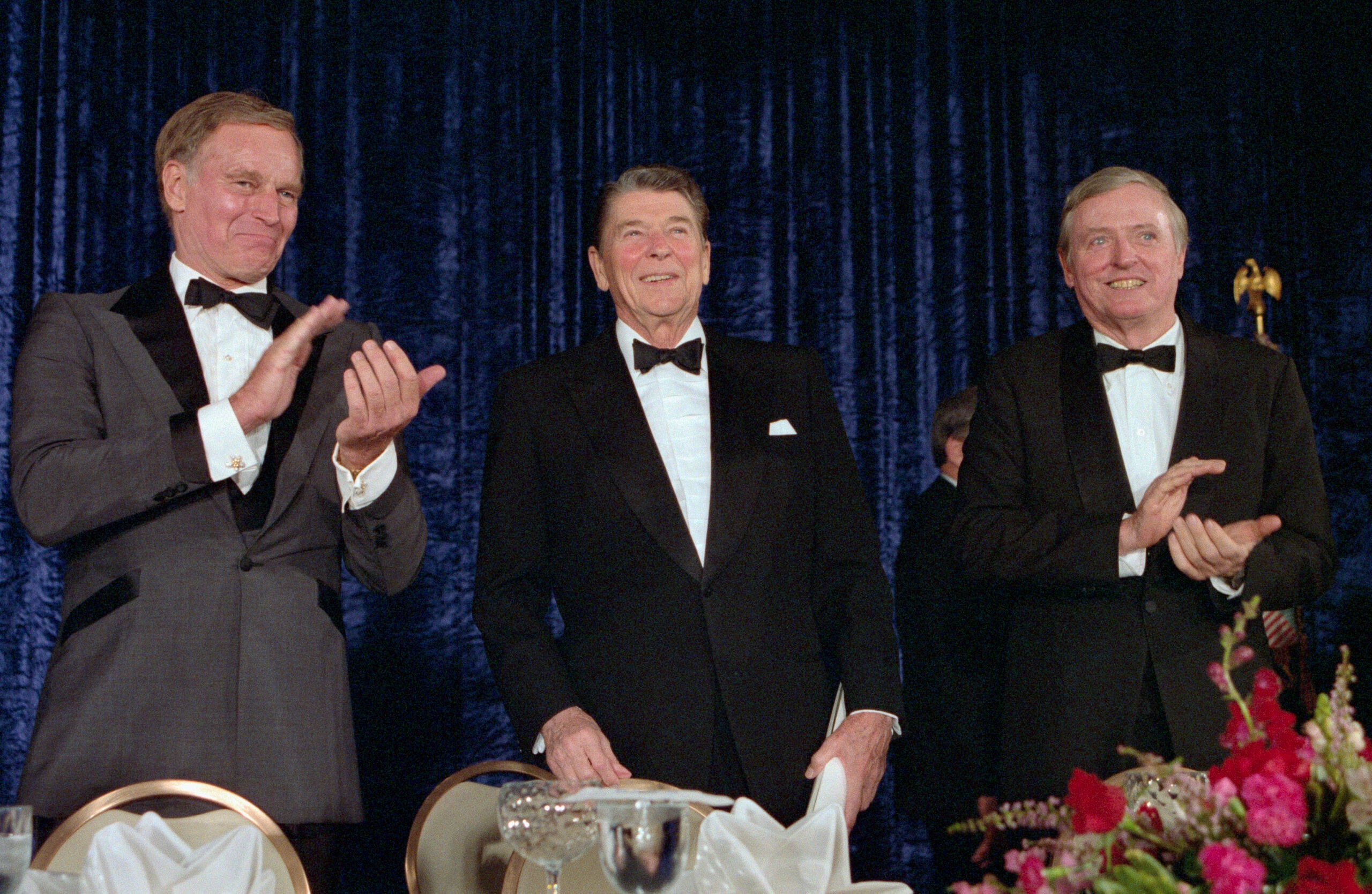 Three men in tuxedos stand together at a formal event. Two are clapping while one smiles at the camera. They are in front of a dark blue curtain with a banquet table and floral arrangement in the foreground.