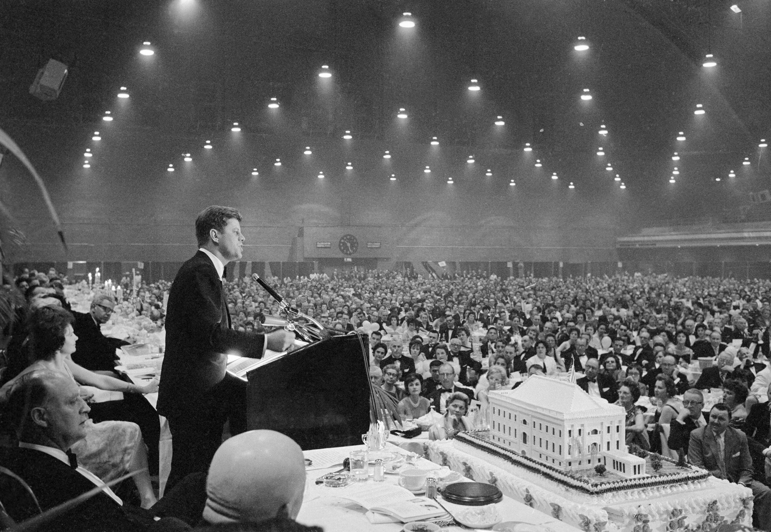 Vintage photos of meals. A man in a suit speaks at a podium to a large audience in a banquet hall; a detailed building-shaped cake sits on the table in front.
