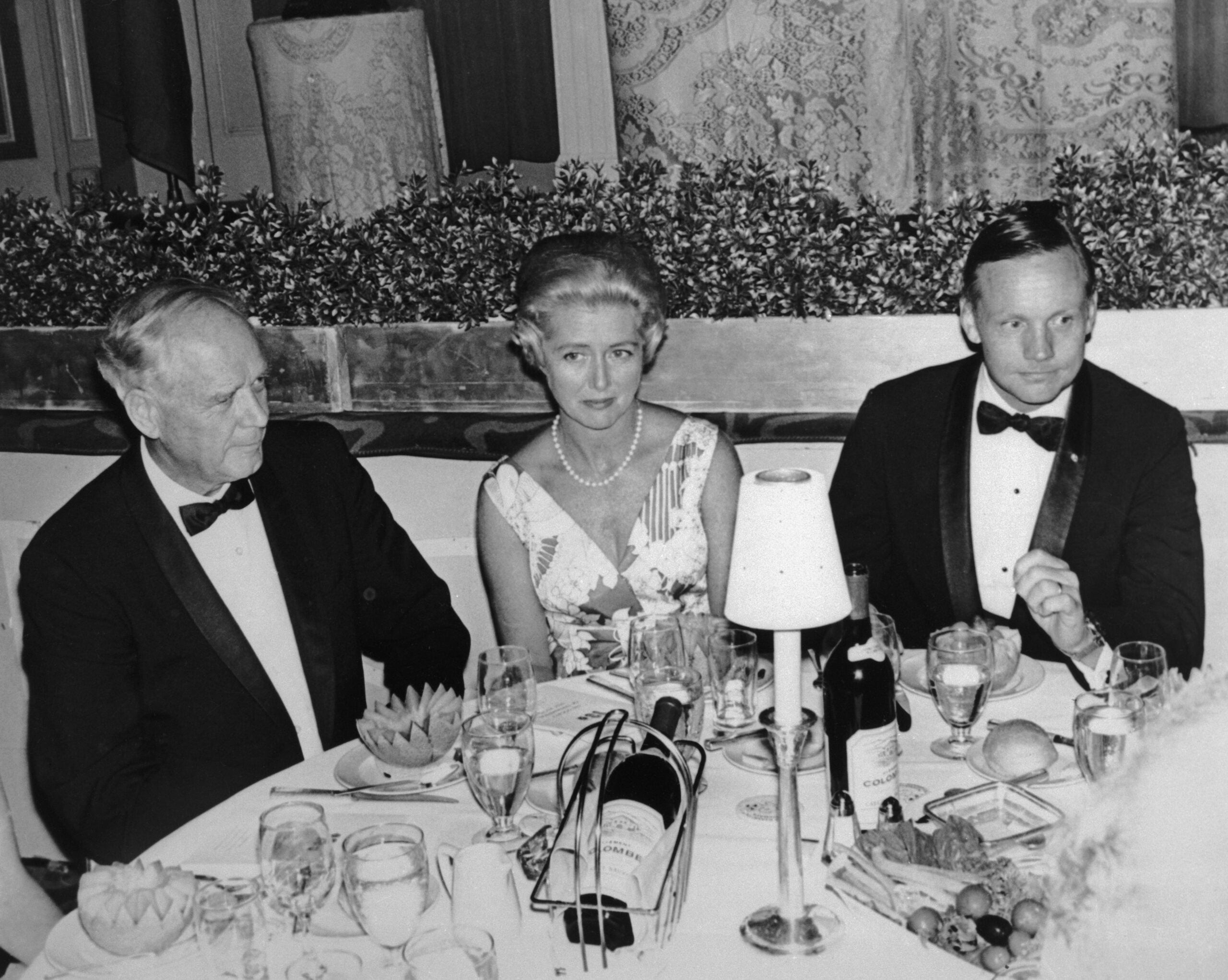 Three people in formal attire sit at a round dinner table set with wine, glasses, plates, and a table lamp, at an elegant event. The woman in the center looks at the camera, while the two men look elsewhere.