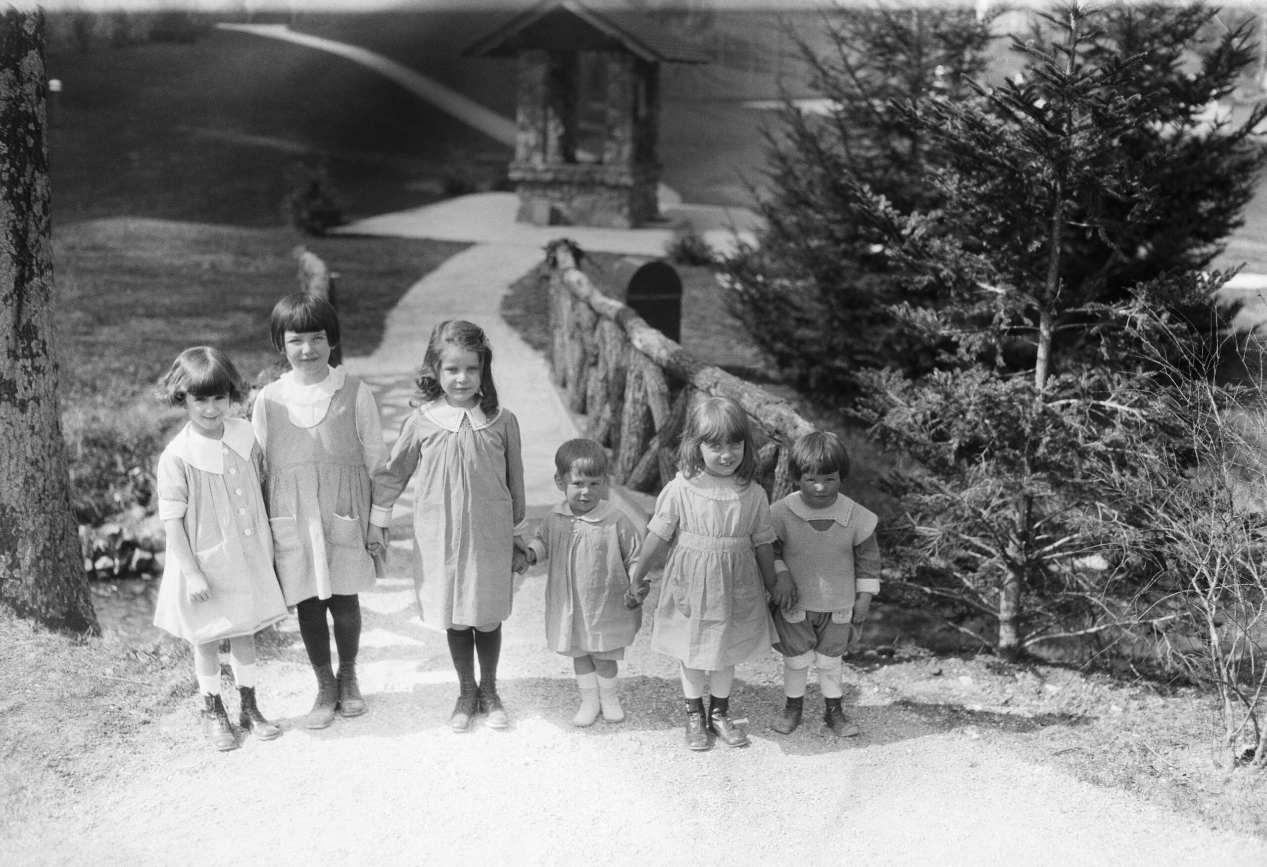 Six young children in vintage clothing stand in a line outdoors near a dirt path, a wooden bridge, and a well, with trees and grassy hills in the background, smiling and holding hands.