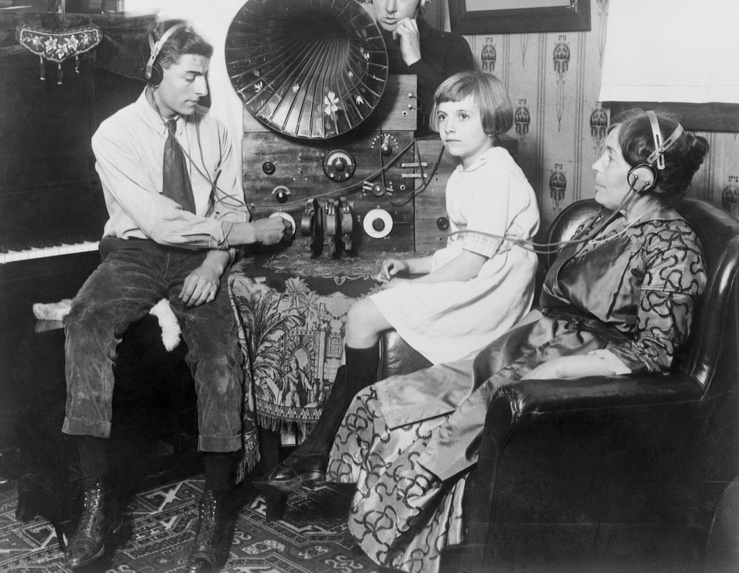 Three people sit around a vintage radio, wearing headphones. A young girl sits on a chair, a man adjusts controls, and a woman sits on an armchair. The scene appears to be from the early 20th century.