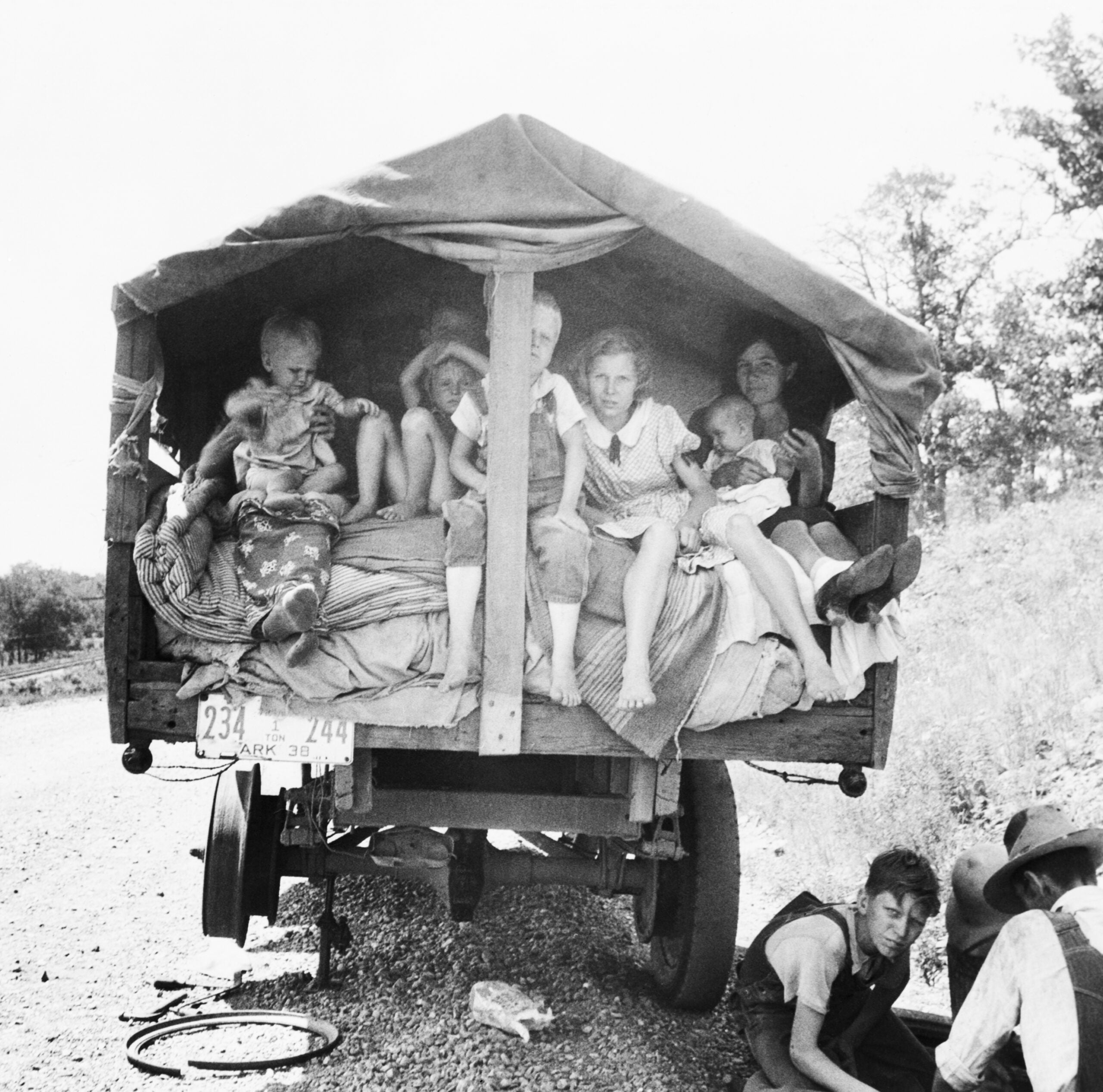 A family sits inside a covered wagon on a rural dirt road. Children and adults rest closely together, with bedding and belongings around them. Two people sit or crouch beside the wagon on the ground.