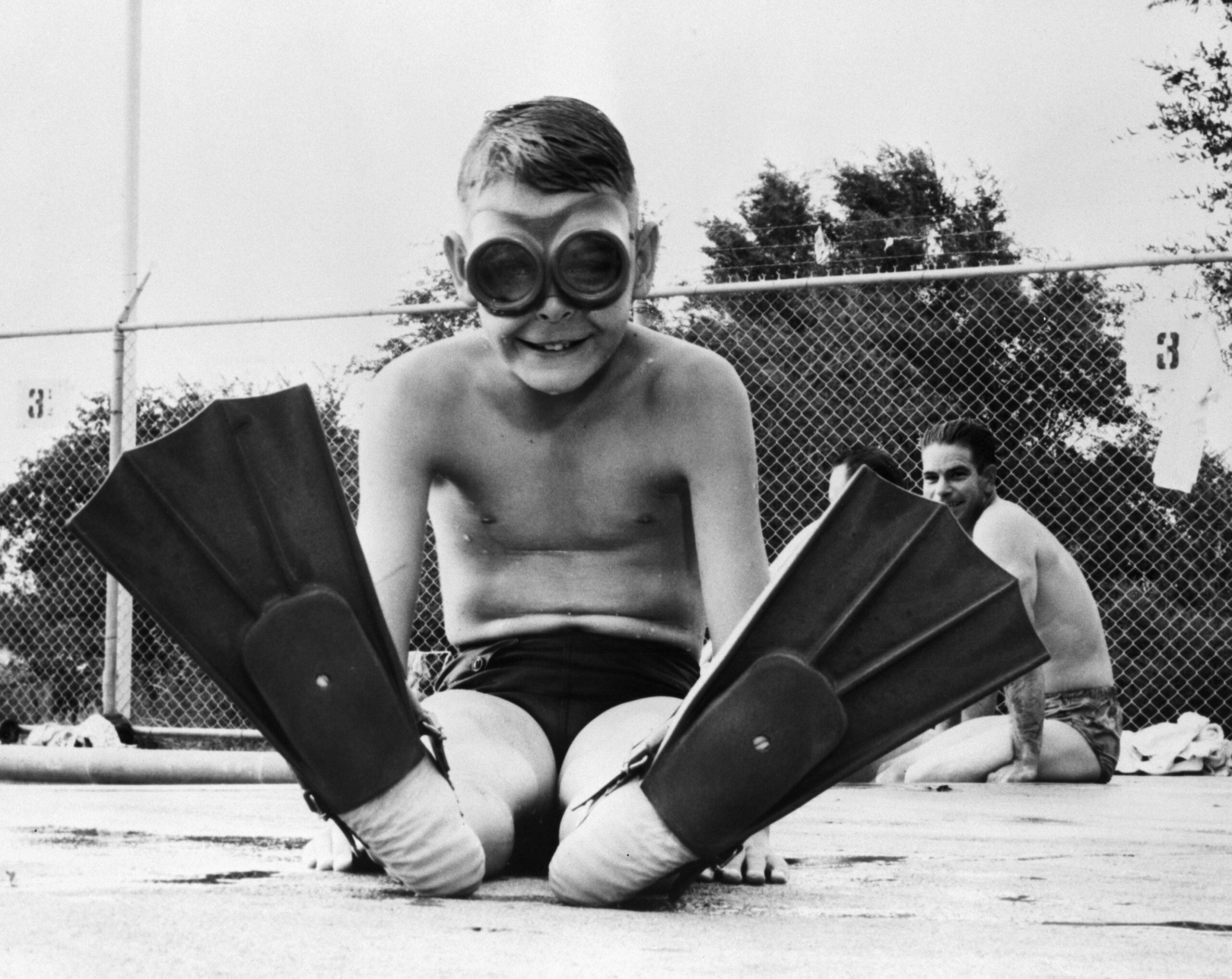 A boy in swim trunks sits poolside wearing large swim goggles and flippers, smiling at the camera. Another person is seated in the background near a chain-link fence.