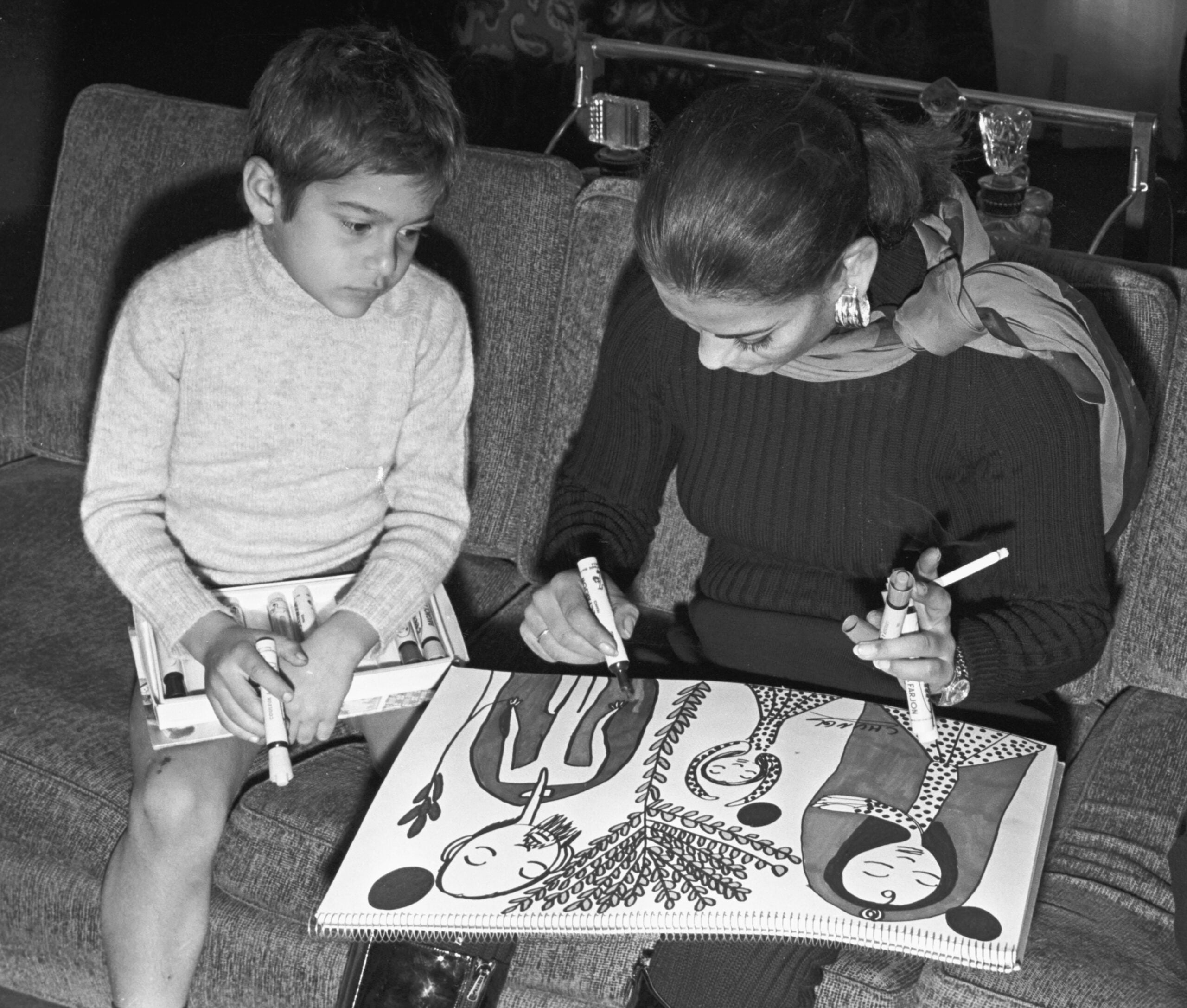 A woman and a young boy sit on a couch, drawing together on a large sketchpad. The woman colors a detailed illustration while the boy observes, holding markers and a coloring book.