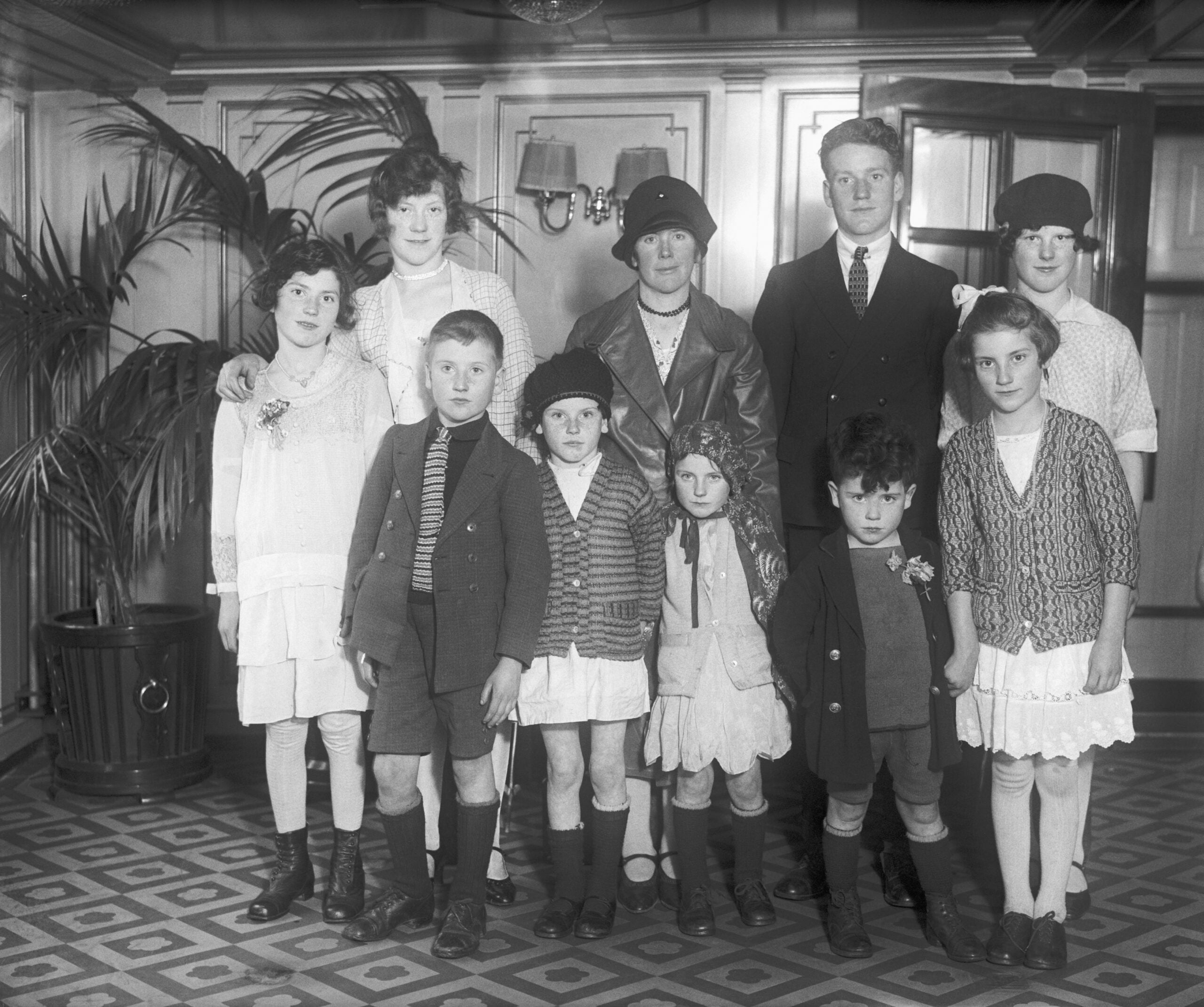 A black and white photo of eight children and two adults in vintage clothing, standing indoors on a patterned floor with plants and wood-paneled walls in the background.