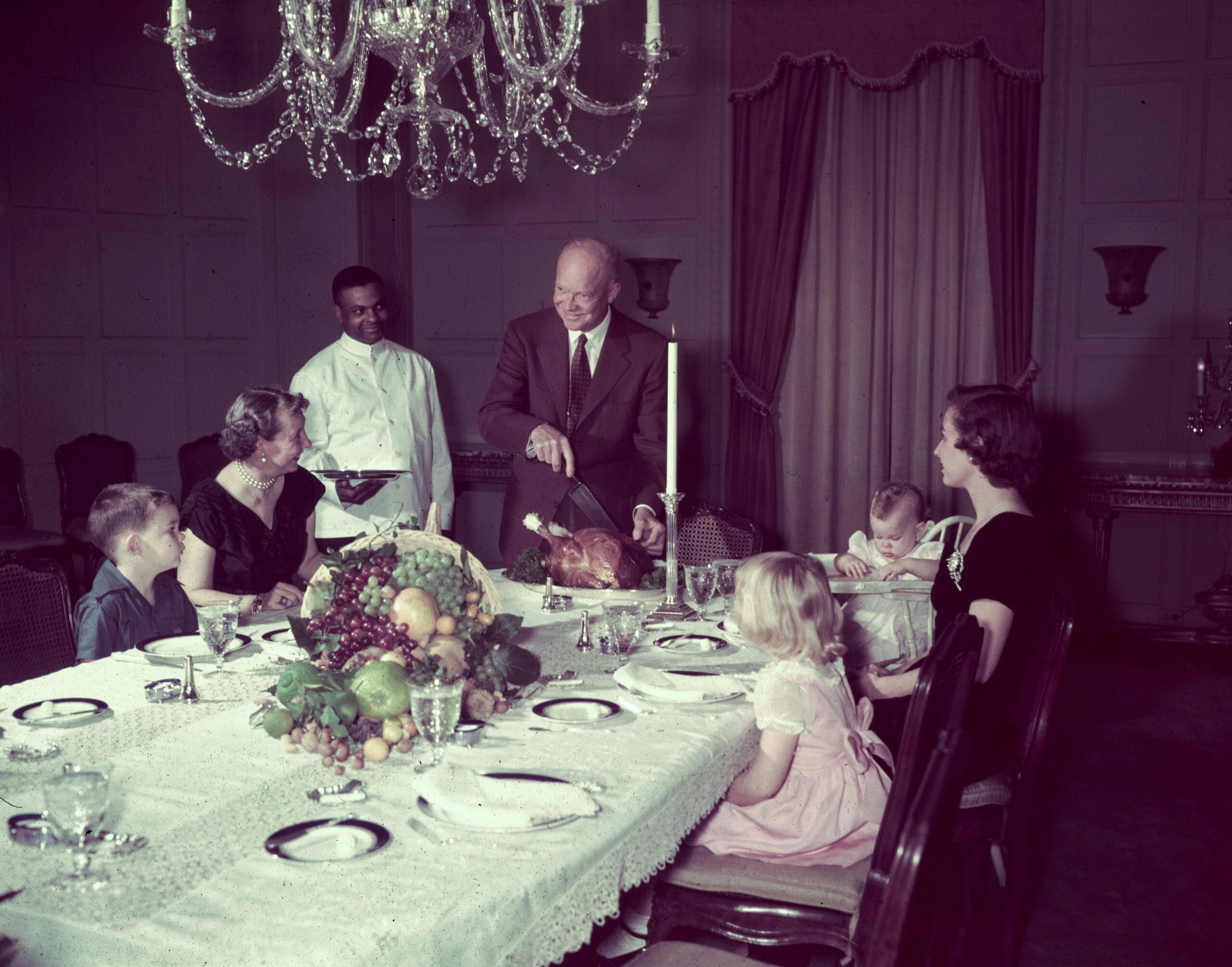 A man carves a turkey at a dining table set with food and surrounded by family, with a waiter standing nearby. The room is elegant, featuring a chandelier and curtains.