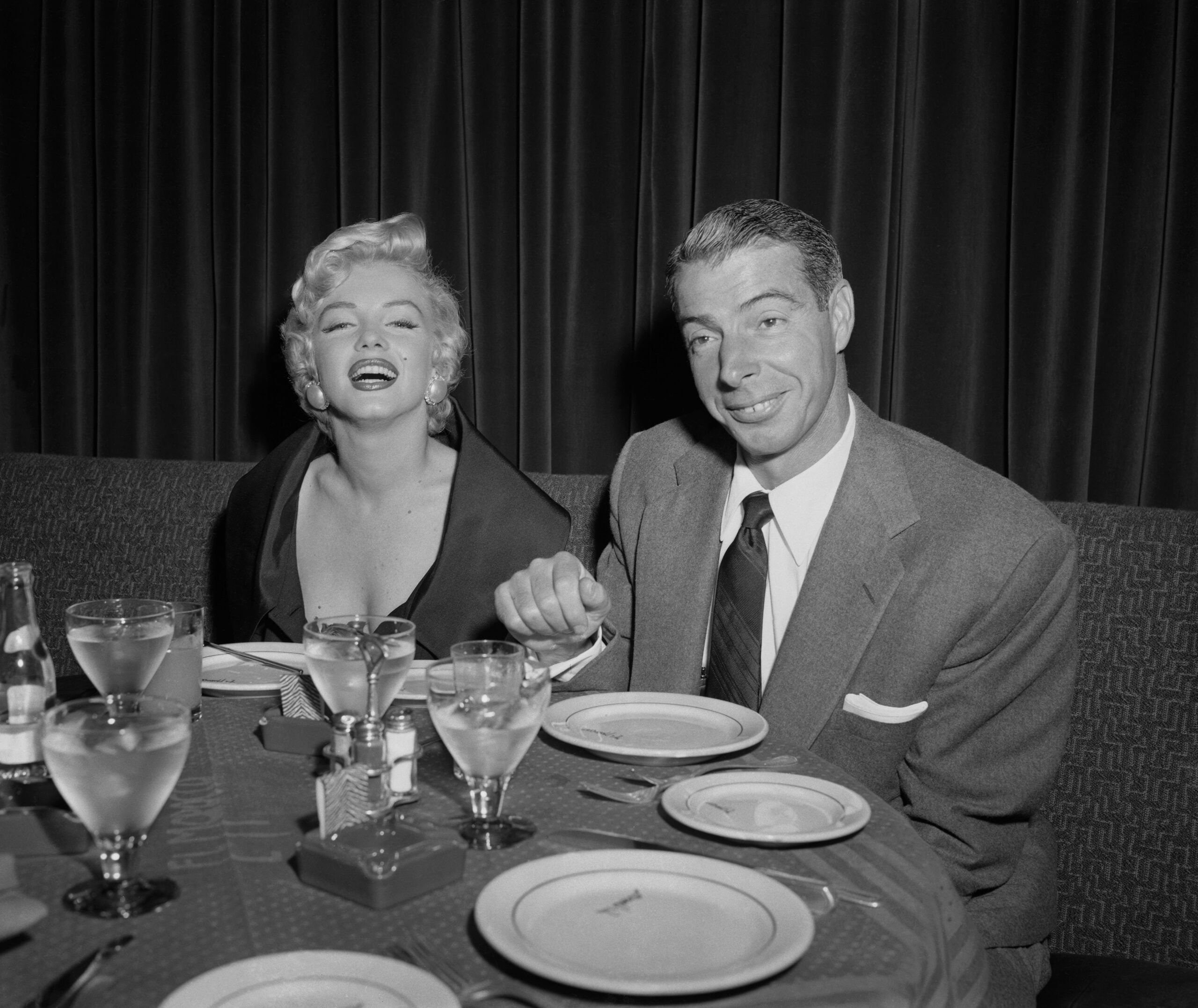 A woman in a dark dress and a man in a suit sit at a restaurant table, smiling and holding hands. There are plates, glasses, and silverware on the table, with a dark curtain in the background.