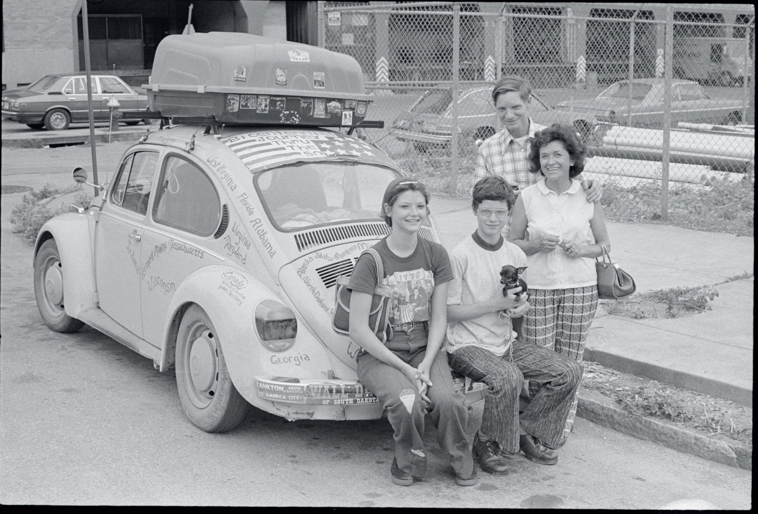 Four people and a dog pose by a Volkswagen Beetle covered in writing and stickers, with camping gear on top. There’s a fence, building, and parked cars in the background.