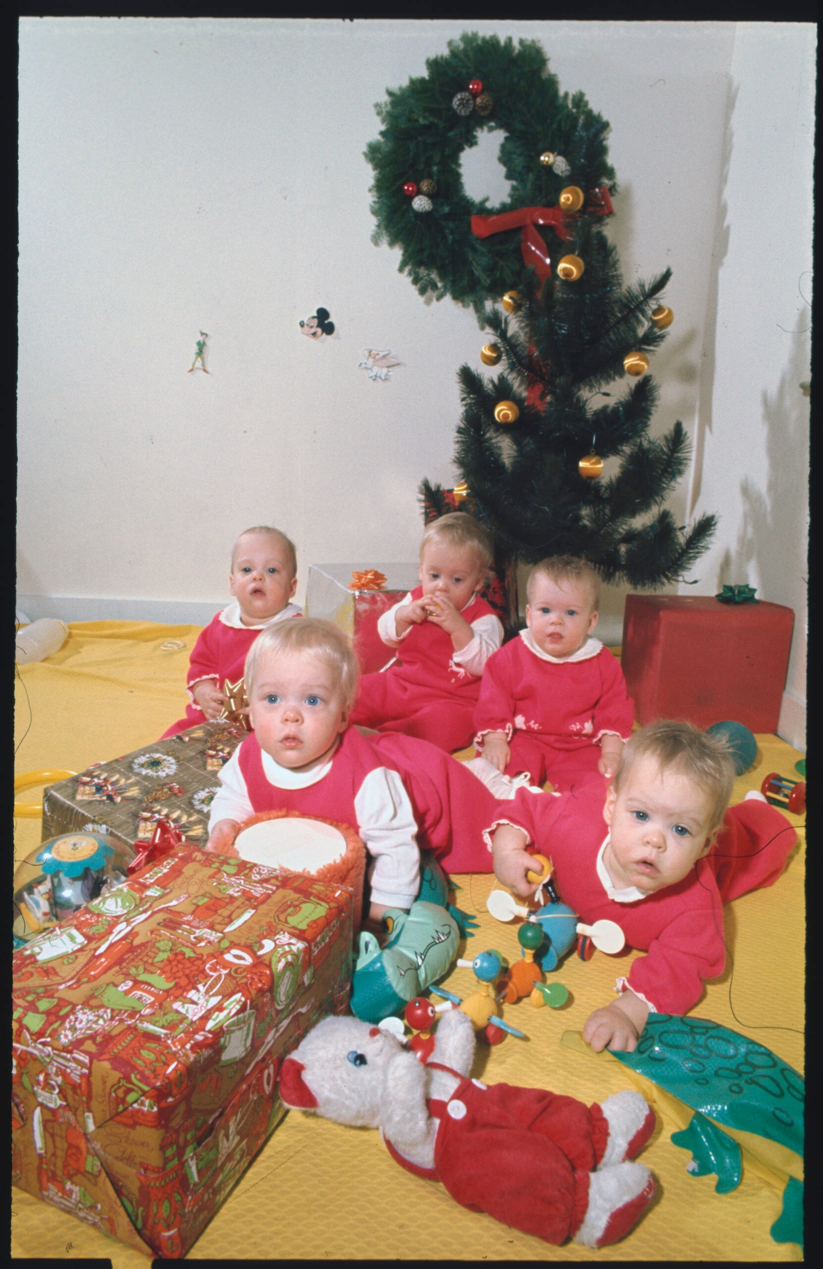 Five babies in matching red outfits sit and crawl on a yellow floor in front of a small decorated Christmas tree, surrounded by wrapped gifts and toys. The scene is festive and brightly lit.