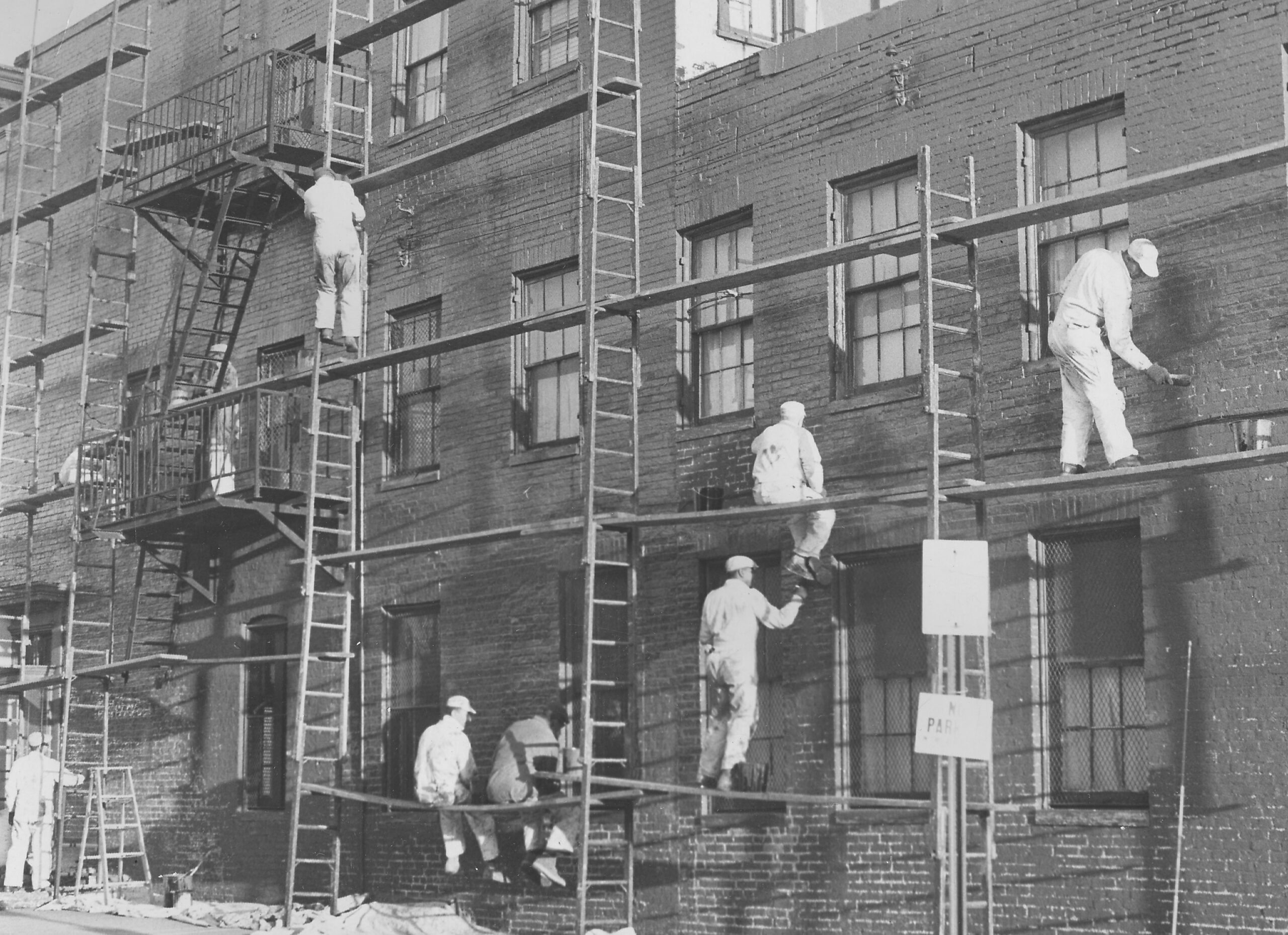 Six workers in white uniforms paint a brick building using ladders and scaffolding. The building has fire escapes and multiple windows. A sign and some equipment are visible at street level.