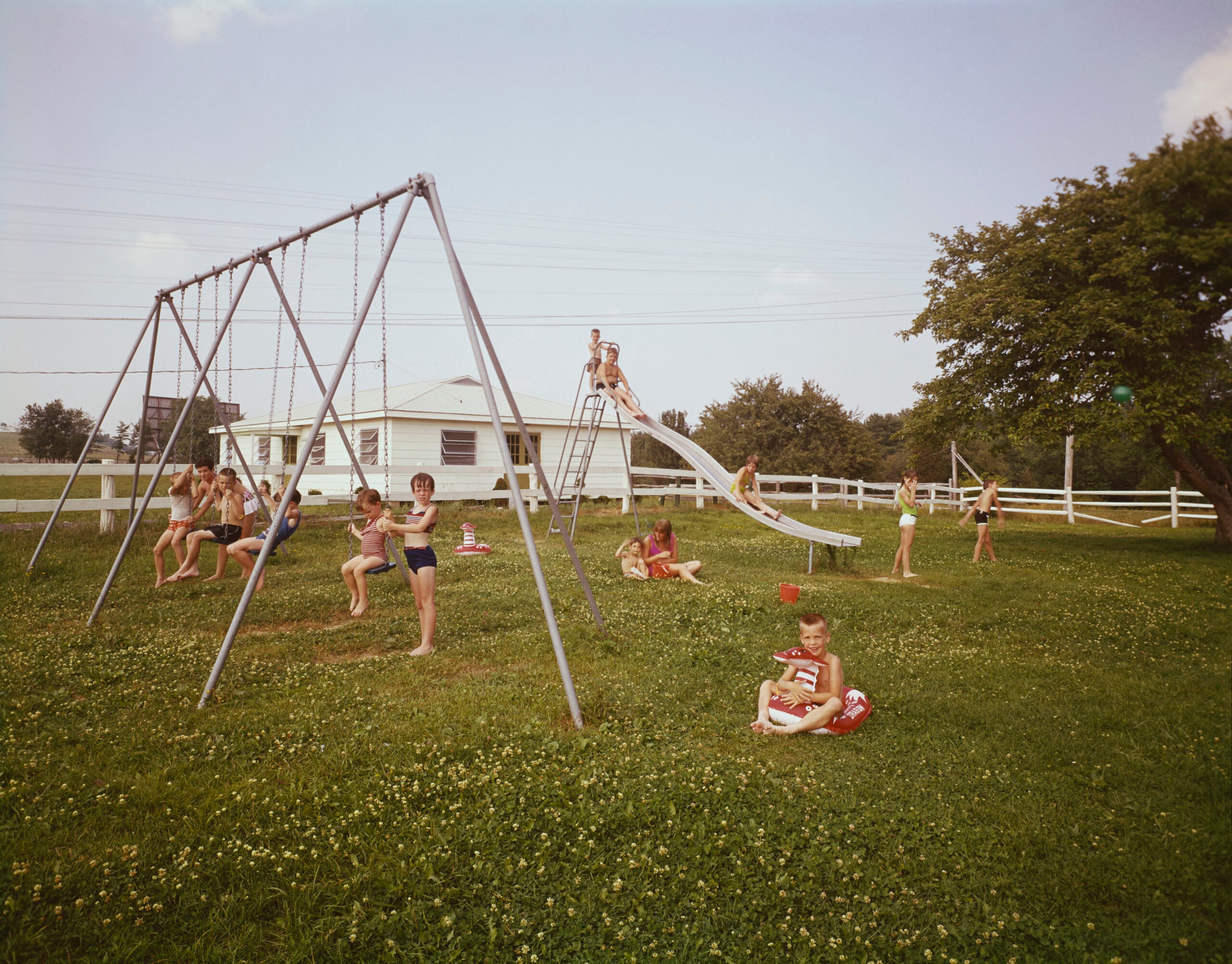 Children play outdoors on swings, a slide, and in the grass near a white fence and house on a sunny day. Some kids swing, others climb or sit, and trees border the yard.