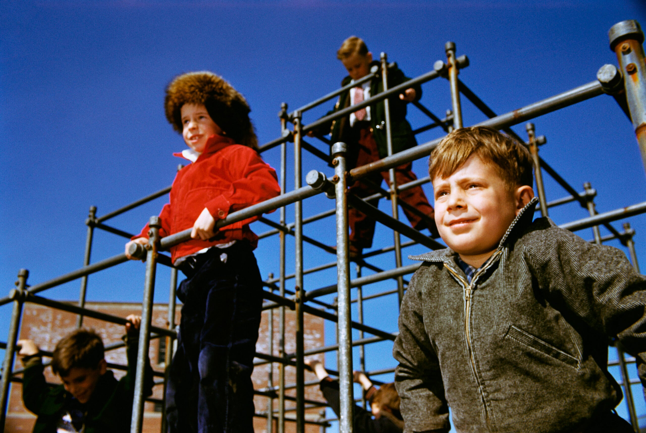 Children play on a metal jungle gym under a clear blue sky, wearing warm jackets and smiling. Some are climbing, while others stand or sit on the structure, enjoying a sunny day outdoors.