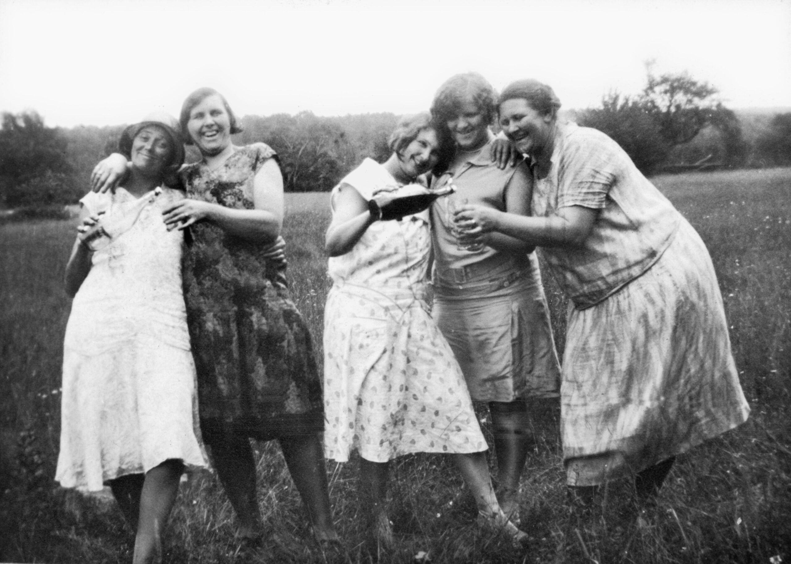 Five women stand together in a grassy field, smiling and laughing, wearing vintage dresses—a snapshot of working class families in the 1920s and 30s. The photo is black and white and appears to be from the early 20th century. Trees are visible in the background.
