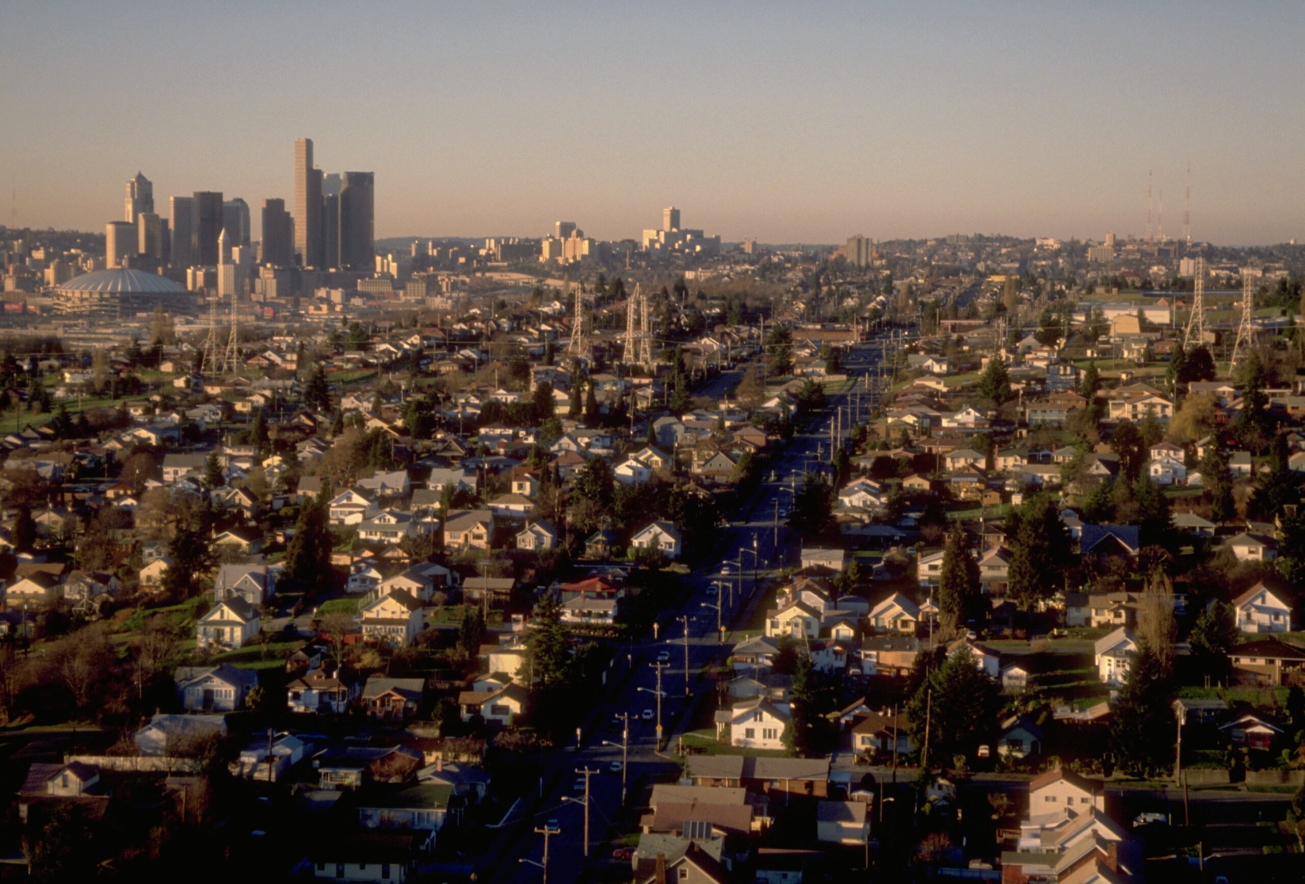 A city skyline in the distance with tall buildings, while a large residential neighborhood with houses, trees, and streets spreads out in the foreground under a clear sky.