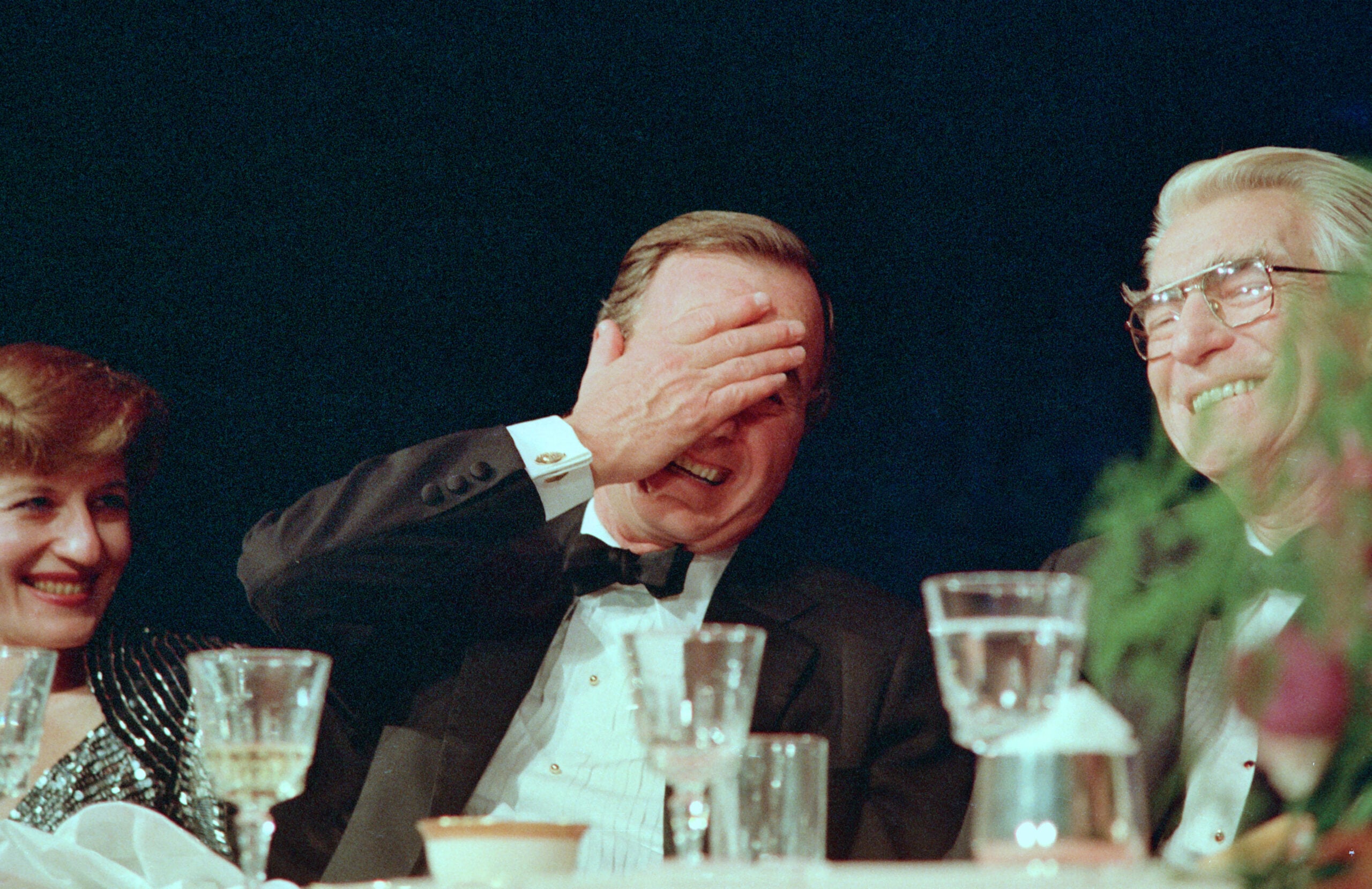 A man in a tuxedo laughs and covers his face with his hand while seated at a table with two other smiling people at a formal event. Several glasses and plates are visible on the table.