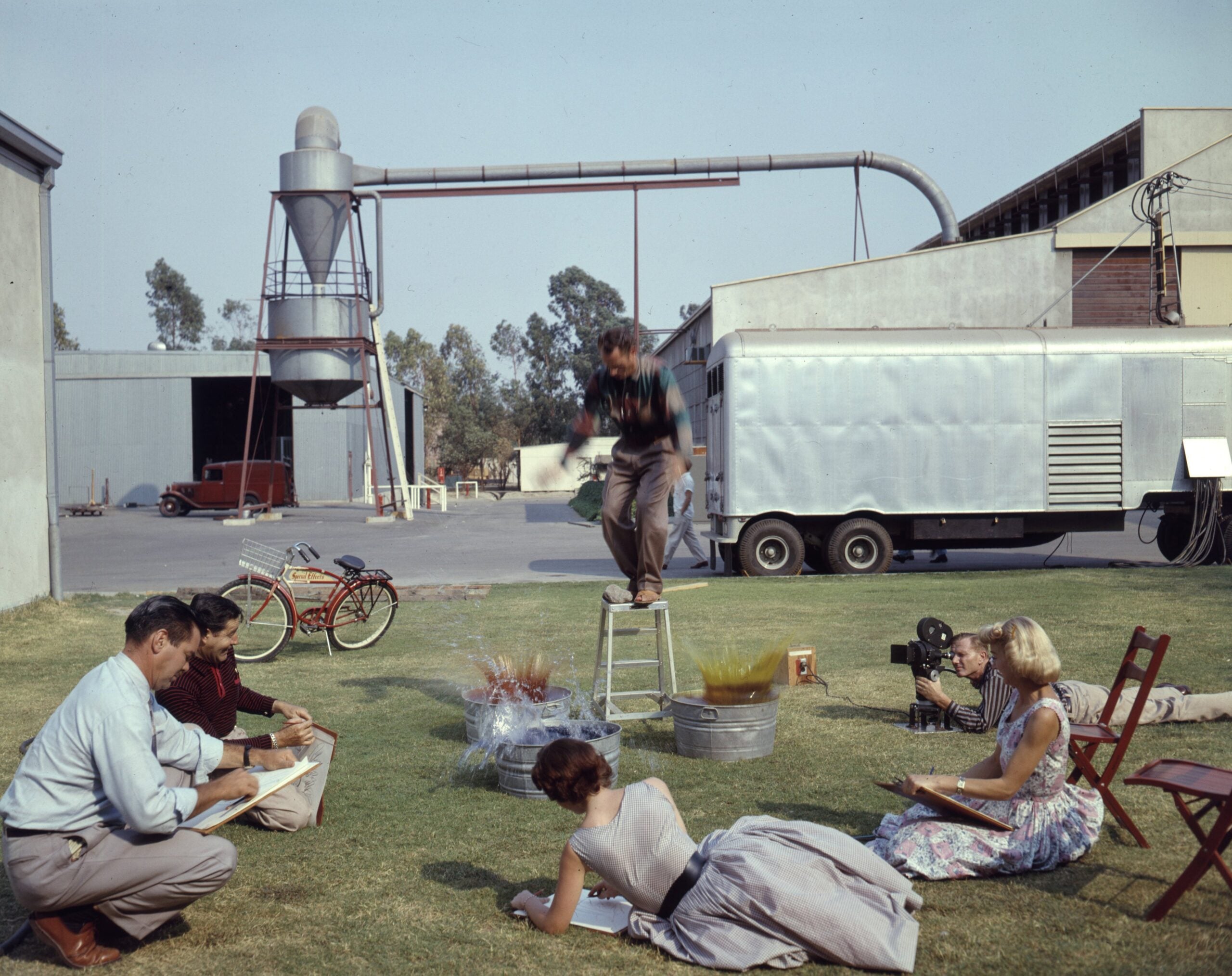 A group of people sketch outdoors as a person stands on a stool, posing. Industrial buildings, a bicycle, and a trailer are visible in the background.