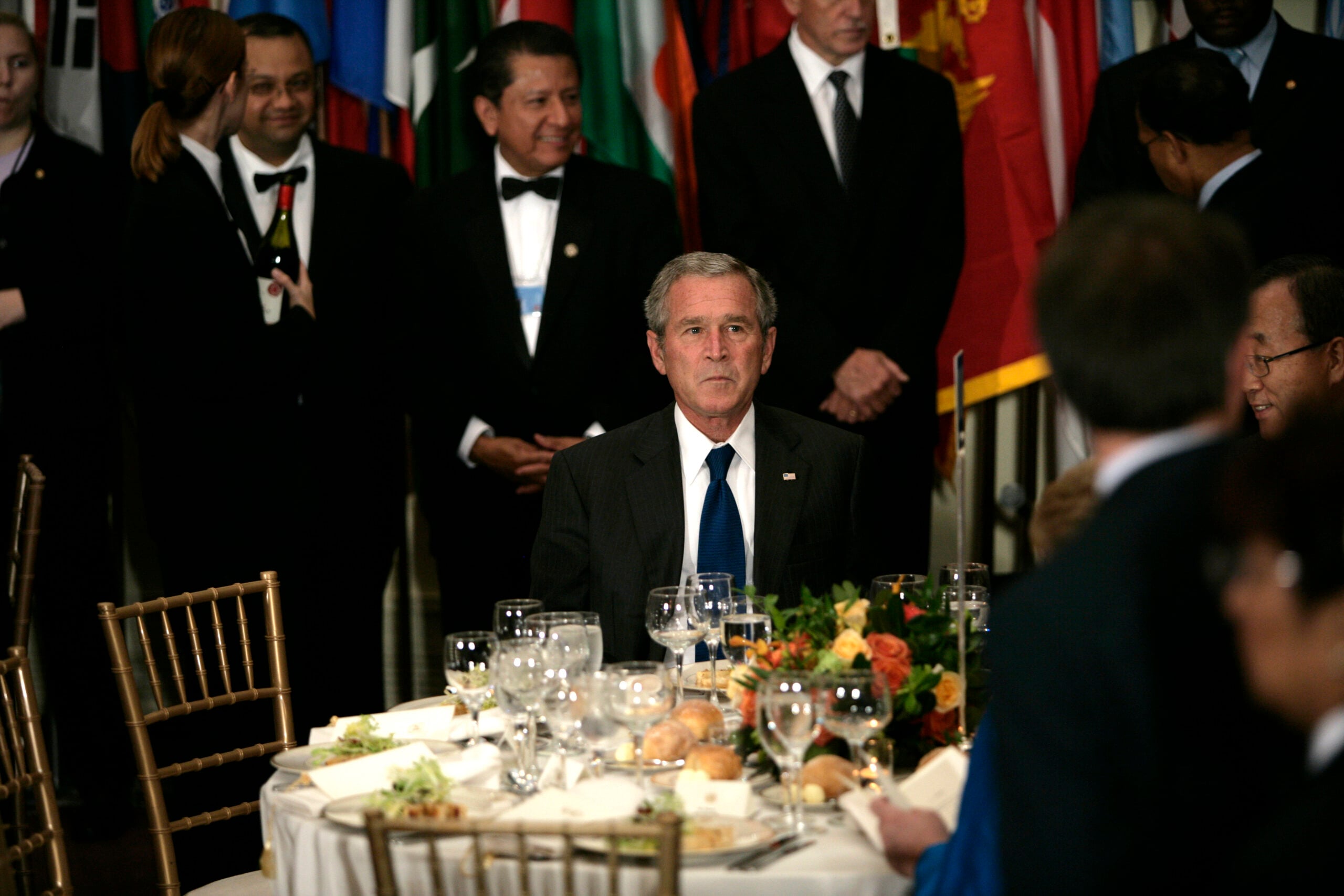 A man in a suit sits at a formal dinner table with empty plates and glasses, surrounded by other people in suits, with international flags in the background.