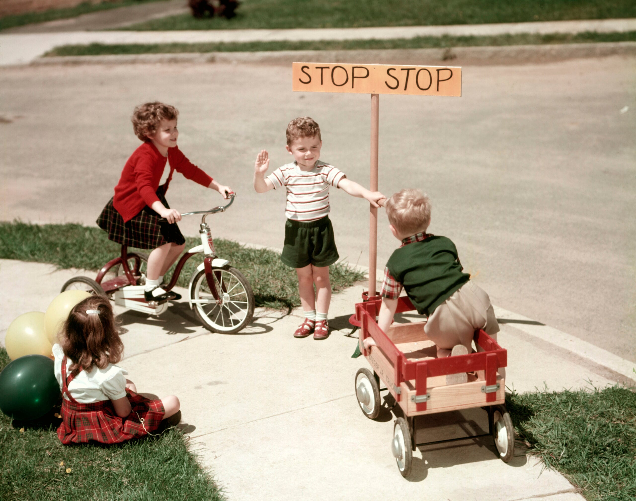 Four children play outdoors; one rides a tricycle, one pulls a wagon, one holds up a hand by a “STOP STOP” sign, and one sits on the grass with balloons. They are near a sidewalk on a sunny day.