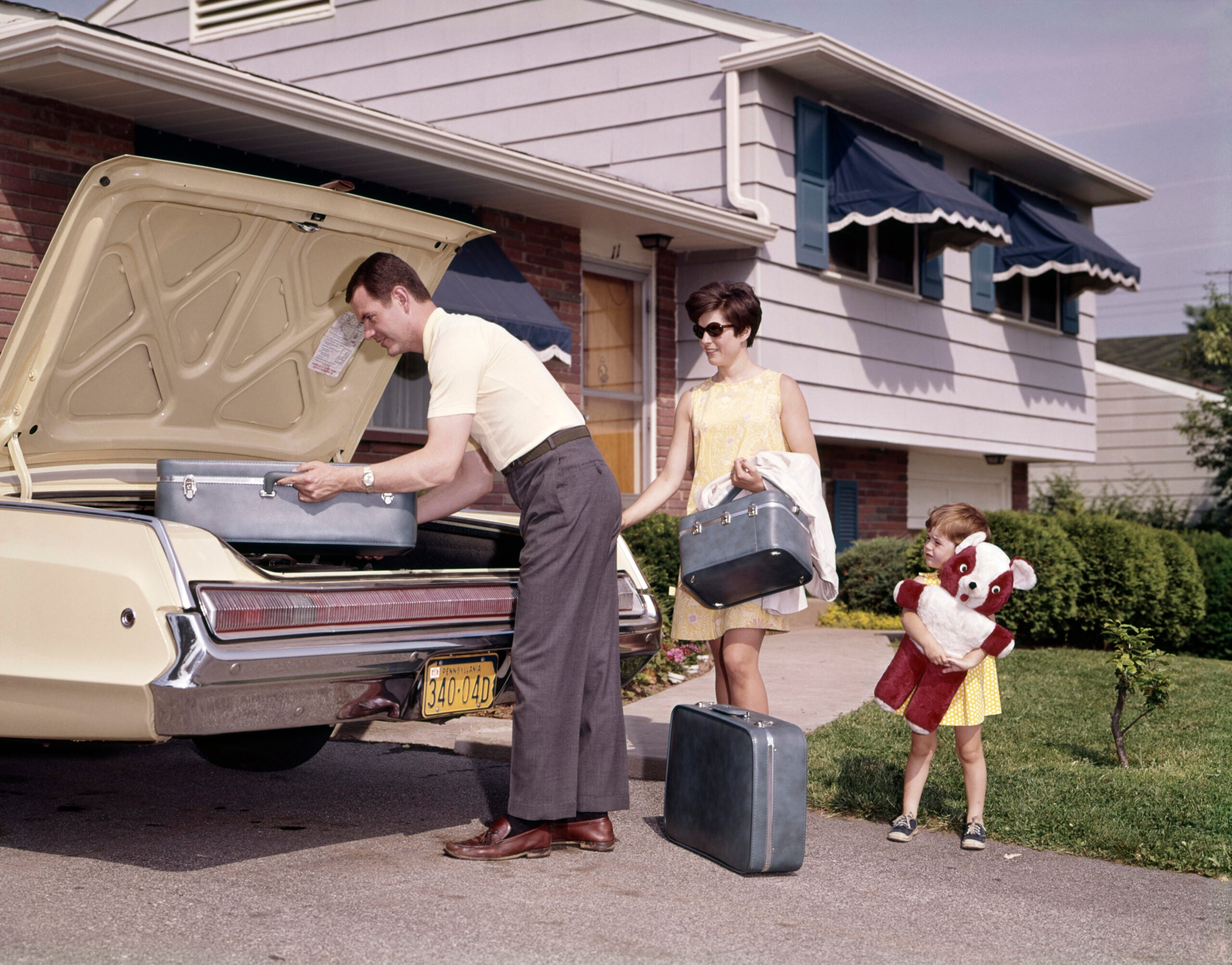 A man loads suitcases into a car trunk while a woman and a young girl, holding a large stuffed animal, stand nearby in front of a suburban house. It appears they are preparing for a trip.