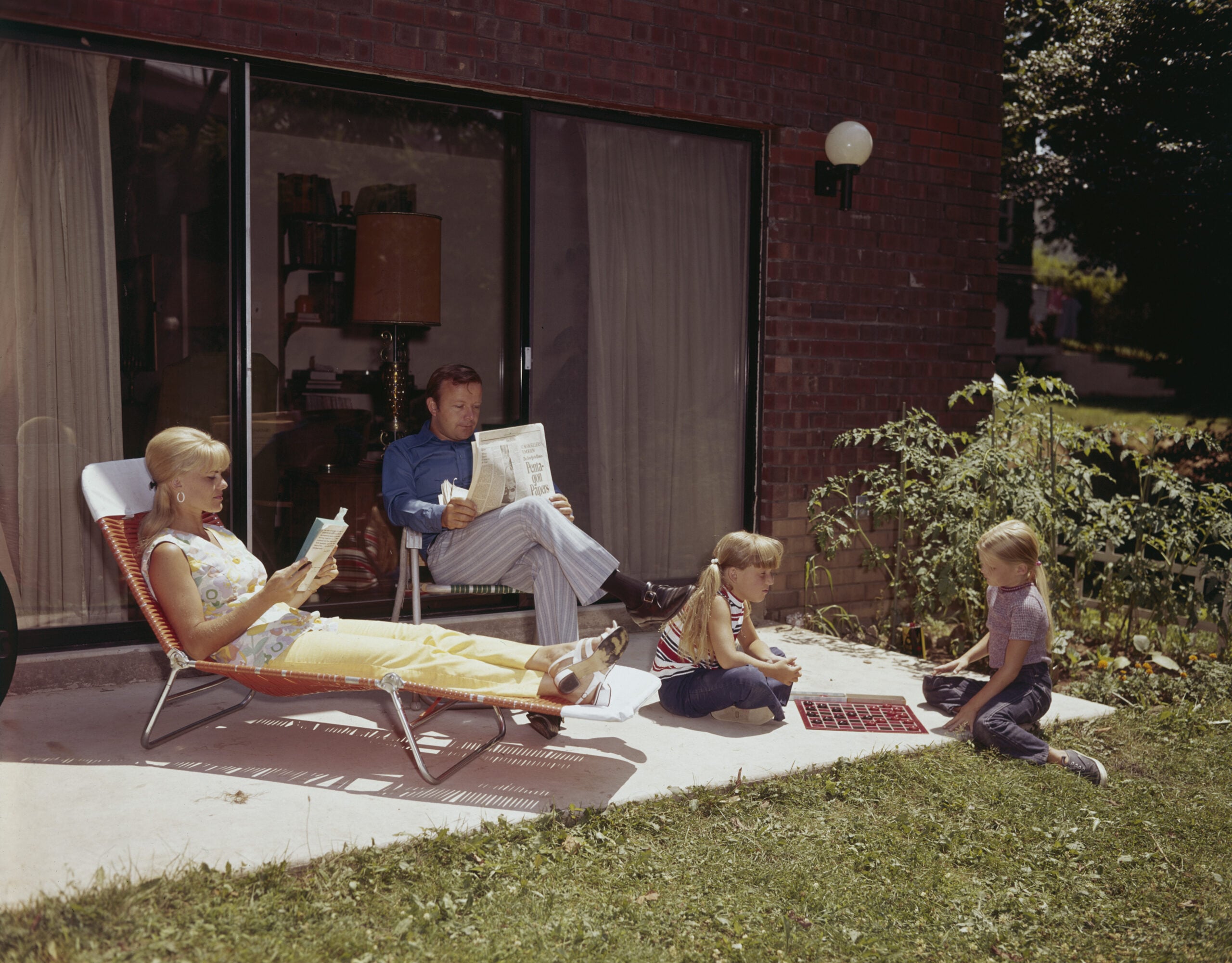 A woman lounges on a chair reading, a man sits reading a newspaper, and two children play a game on the grass outside a brick house with large glass doors.