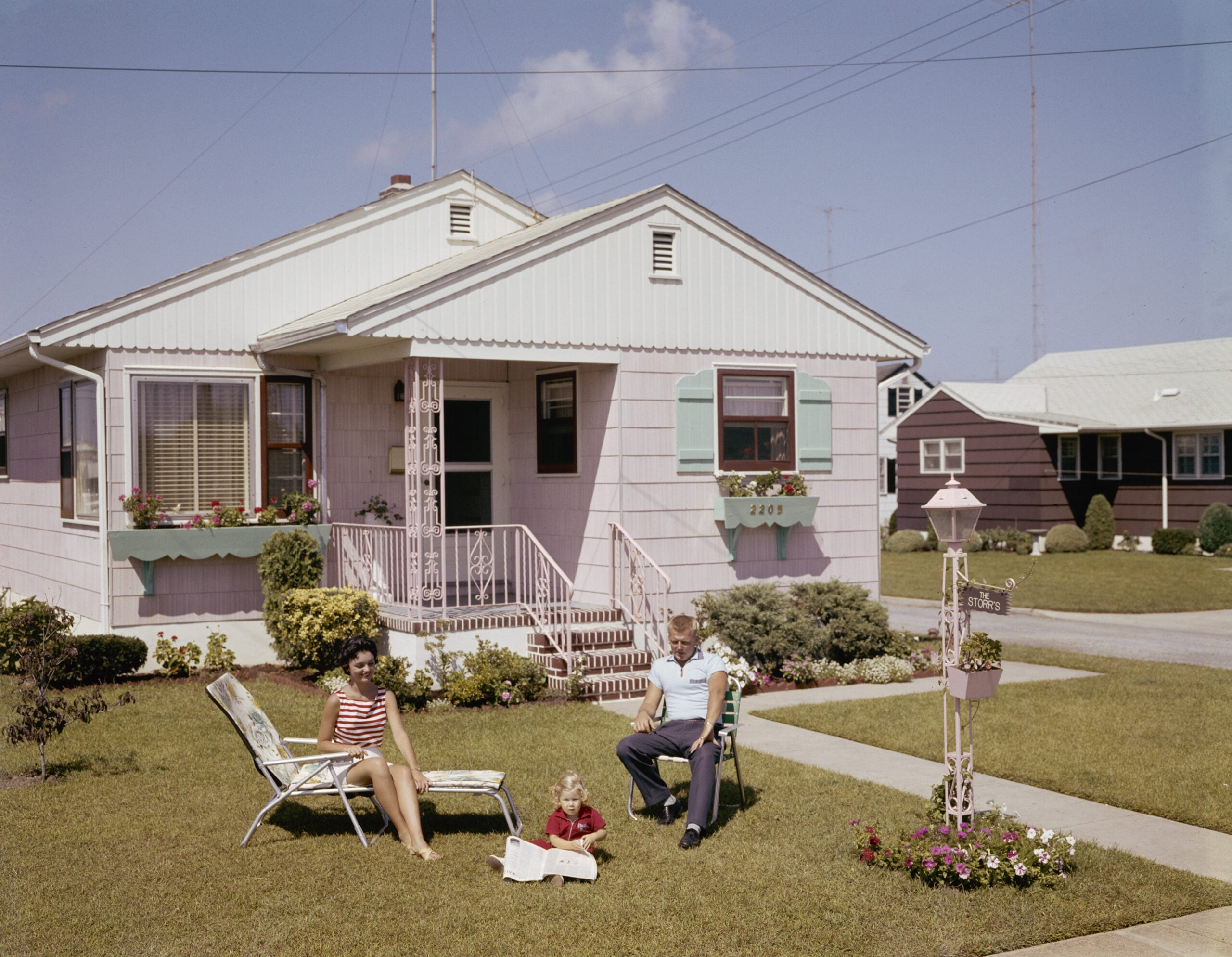 A woman, man, and child relax on lawn chairs outside a light-colored suburban house on a sunny day. The yard is neatly maintained with bushes, flowers, and a birdhouse.
