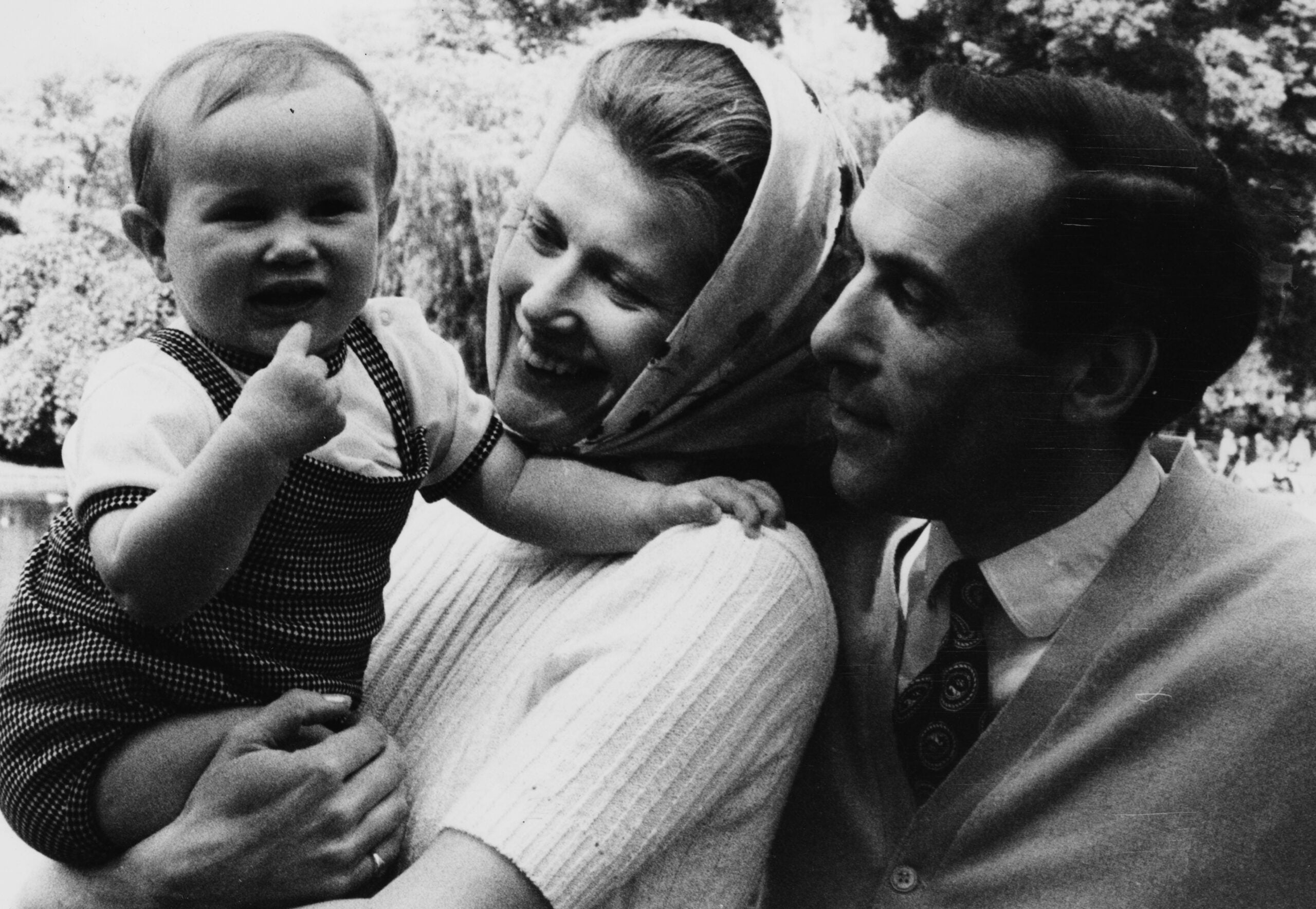 A smiling woman holds a baby who is giving a thumbs-up, while a man stands close beside them outdoors with trees in the background.