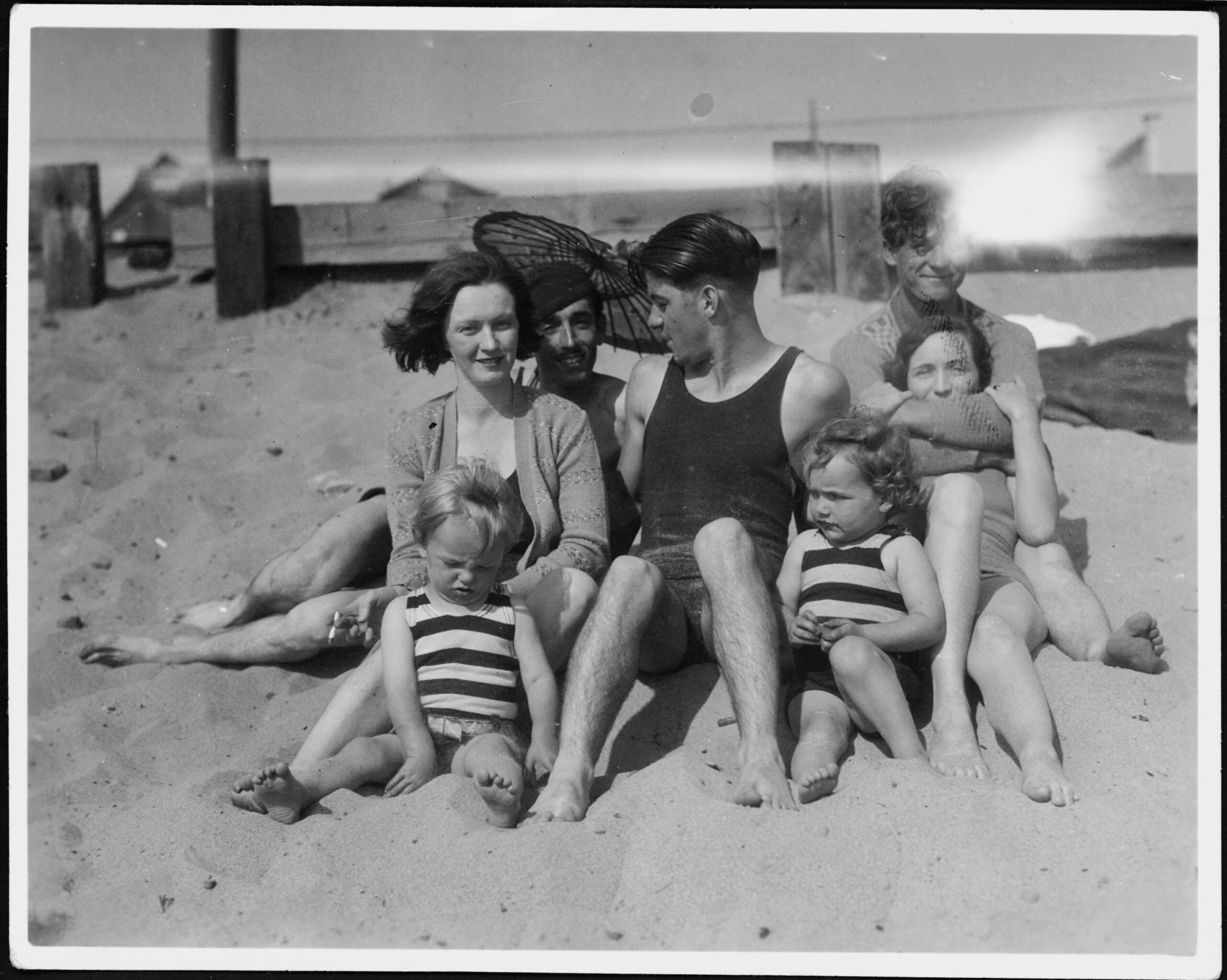 A vintage black-and-white photo of six adults and two small children sitting close together on a sandy beach, wearing swimsuits. Some are smiling, and a wooden structure is visible in the background.