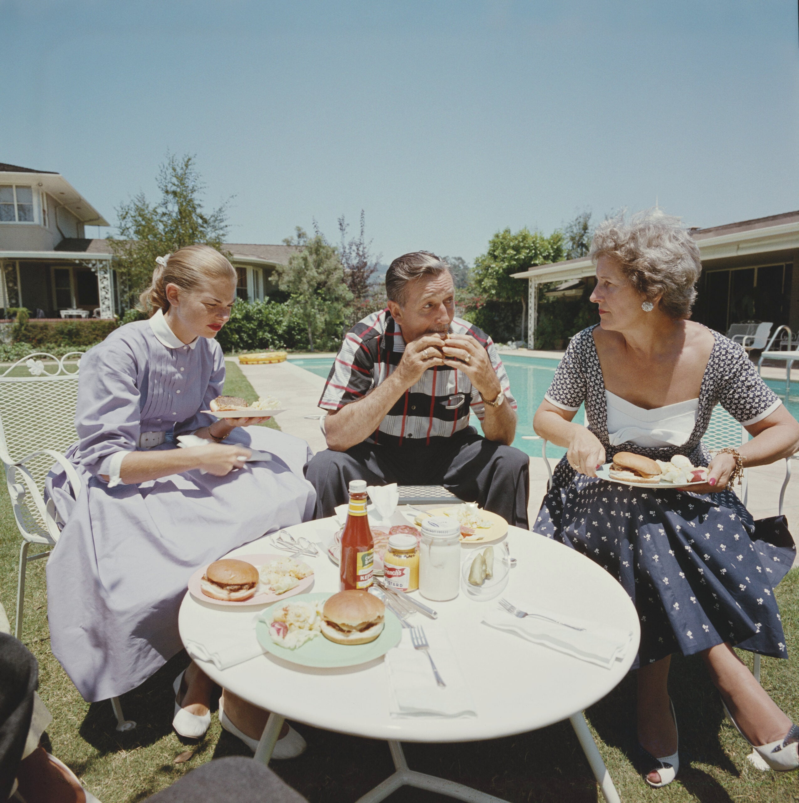 Three people sit at a round table by a pool, eating burgers. Two women and a man are smiling. The table has plates, food, and condiments. Houses and trees are visible in the background under a clear sky.