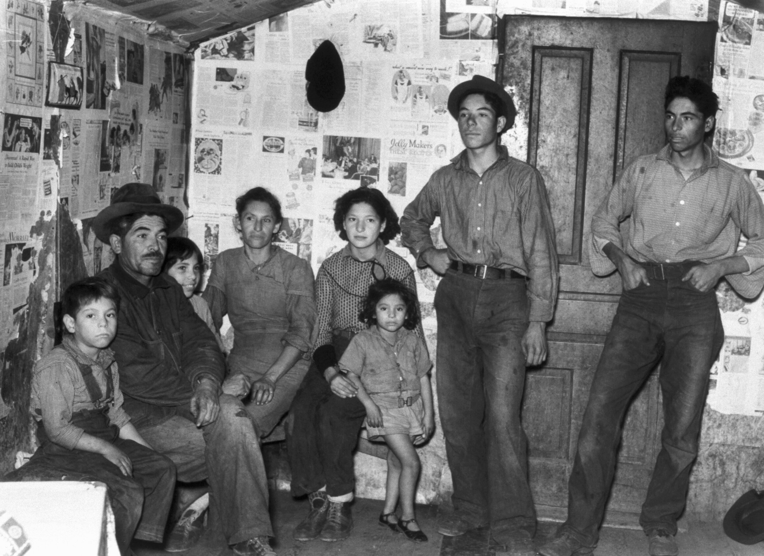 Seven people, including children and adults, sit and stand in a small room with walls covered in newspaper. They wear simple clothing and appear serious, gazing toward the camera.