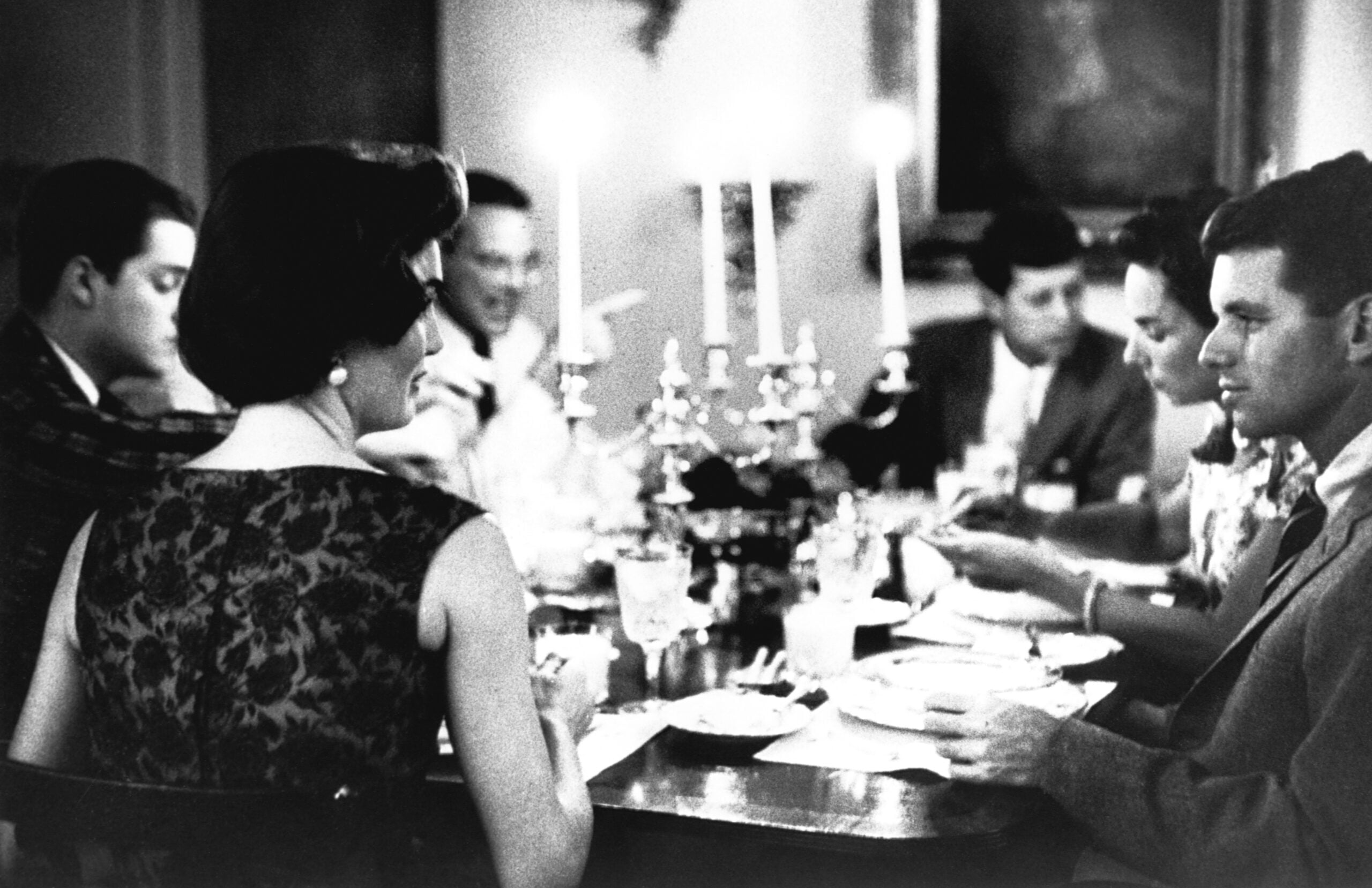 Six people dressed formally sit around a candlelit dinner table, engaged in conversation and eating. The setting is elegant with candlesticks and glassware on the table, and the image is in black and white.